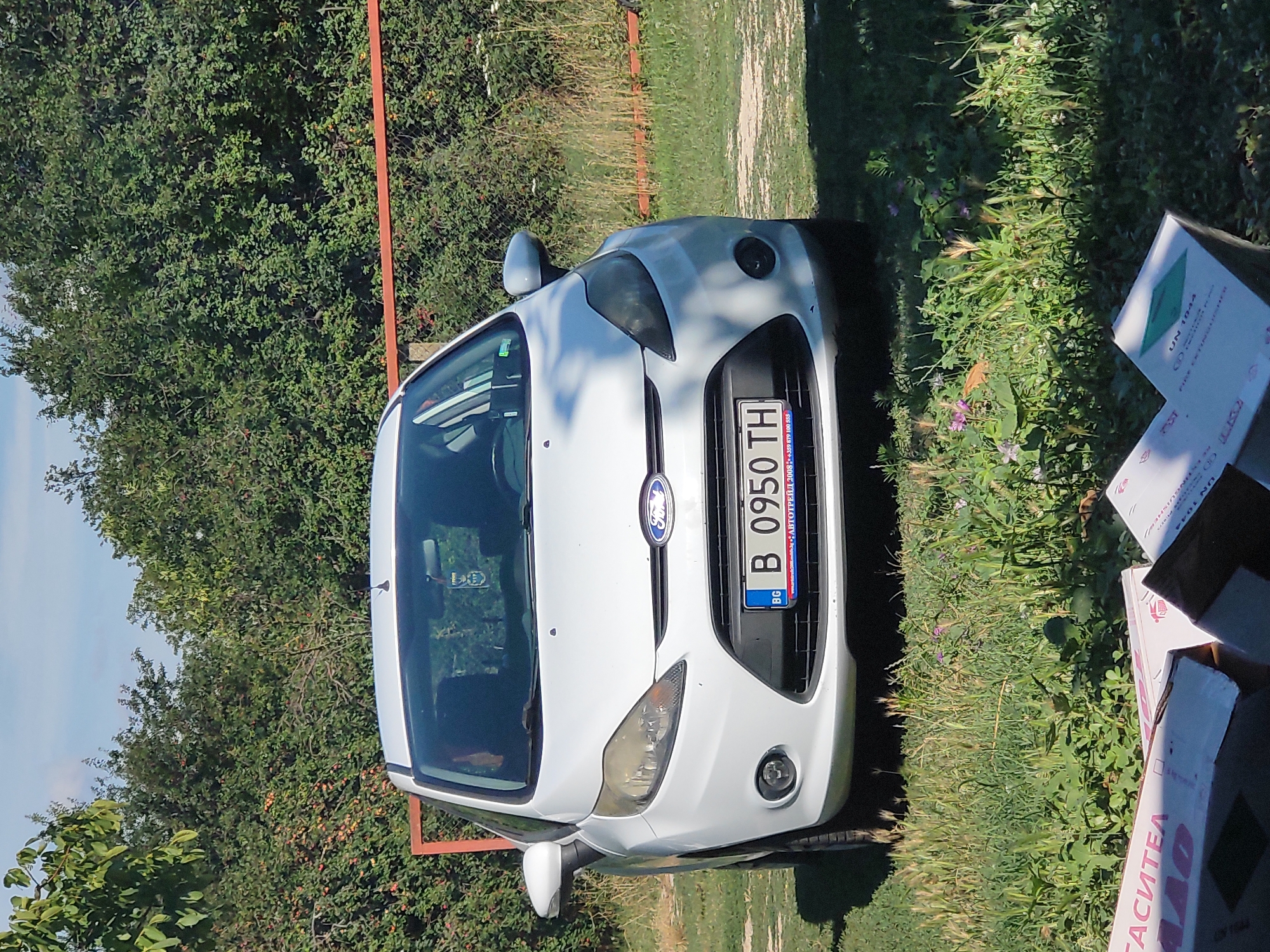A white Ford Fiesta is parked on a grassy area with trees and a red fence in the background. The car's license plate reads "B 0950 TH". In the foreground, several cardboard boxes are visible. The scene appears to be in Kochmar, Bulgaria.