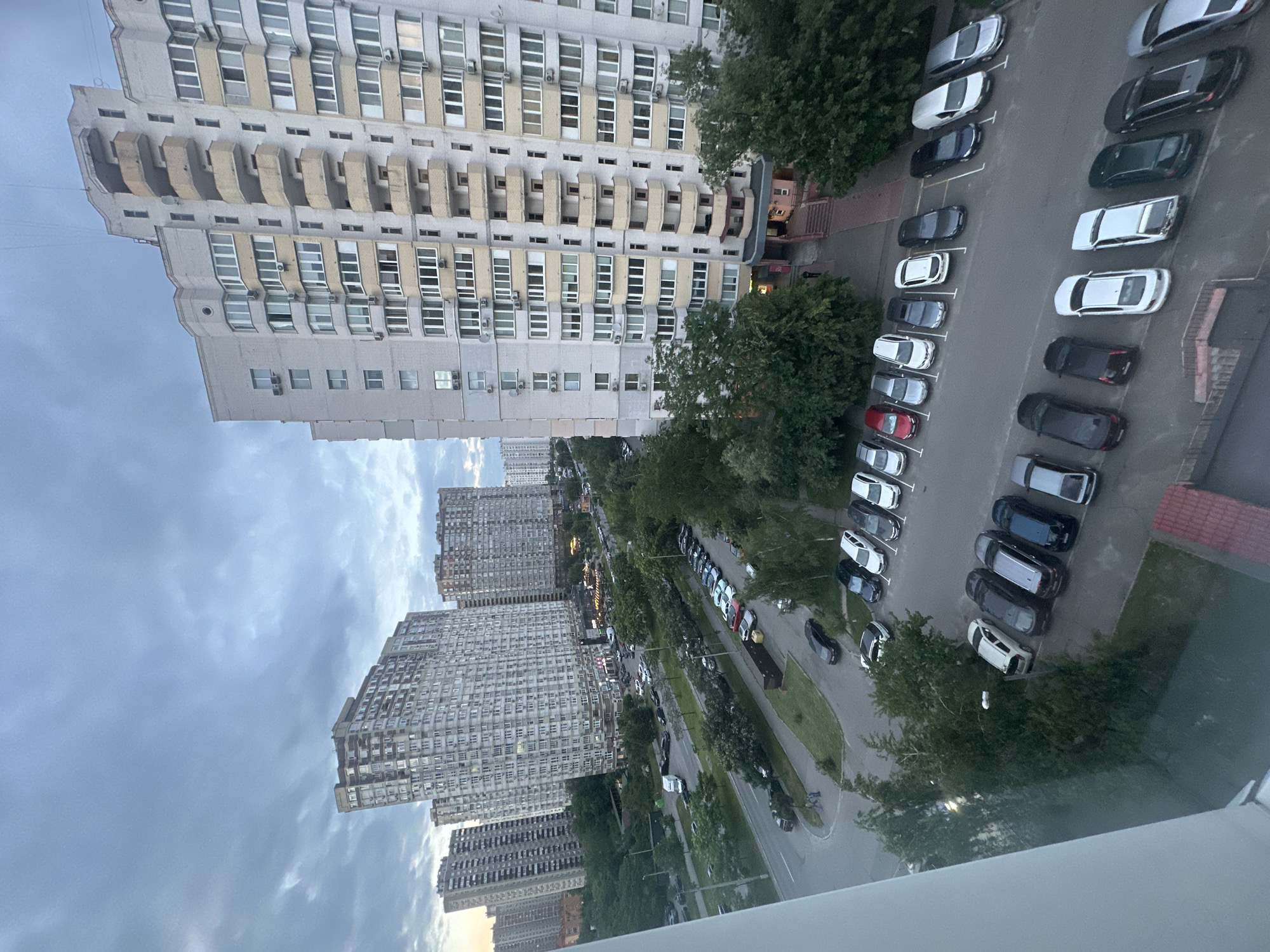 This is a medium-high angle, daylight, medium shot photograph overlooking a residential area in Kyiv, Ukraine. The sky is overcast with heavy grey clouds. In the foreground, the corner of a white building is visible on the left. To the right, a tall, multi-story apartment building with a light-colored facade and numerous balconies is the dominant structure. The building has a modern, block-like design with many windows.
Below the apartment buildings, a wide asphalt road runs horizontally across the middle ground of the image. A variety of cars are parked along the side of the road and in designated parking lots. The cars are of different makes and colors, including white, silver, black, and dark blue. Several trees with dense green foliage line the road and parking areas, creating a somewhat urban park-like setting.
Further in the background, more tall apartment buildings, similar in style to the main one, are visible, extending into the distance. The overall impression is of a densely populated urban residential district. There are no obvious human subjects in action, with only a few figures visible near the entrance of the main apartment building. The scene appears to be calm and everyday.