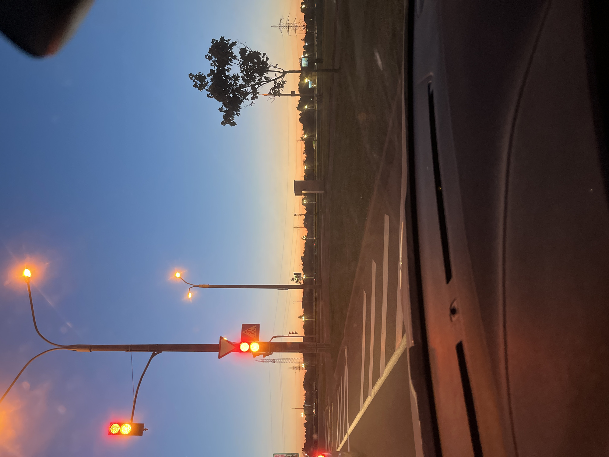 The image depicts a roadside scene at either sunrise or sunset, captured from within a vehicle. The sky dominates the upper portion, showcasing a serene gradient from deep blue at the zenith to warm orange and yellow tones along the horizon.
In the foreground on the left, several illuminated streetlights extend from poles, casting a soft, warm glow. Multiple red traffic lights are clearly visible, indicating that traffic is stopped. A prominent traffic light fixture in the center displays two illuminated red signals. Attached to the same pole are a white triangular sign, possibly a yield sign, and a rectangular sign, but any text on them is unreadable. Further down and to the left, another red traffic light for vehicles is seen, along with a partially visible red pedestrian signal below which some blurry white text, possibly "FONDPETRY," can be faintly discerned.
The right side of the image is occupied by the dark interior of the vehicle, including what appears to be a window frame or car pillar and part of the dashboard. A section of the road with clear white zebra crossing markings is visible on the right.
Along the horizon, the landscape features scattered trees, several illuminated streetlights lining the roadside, and visible power lines. A tall, silhouetted power transmission tower stands in the middle distance against the vibrant sky. The scene suggests an urban or suburban road environment in Minsk, Belarus, during the golden hour, characterized by clear weather and the absence of any visible people.