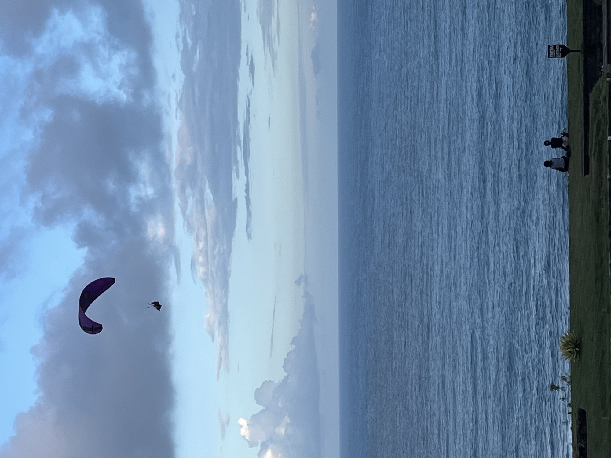 The image depicts a coastal scene. In the foreground, there is a strip of grass and vegetation, with two people standing near a sign. Beyond the grass is a body of water, possibly the ocean, characterized by a flat surface with subtle ripples. In the sky, there are varying cloud formations, ranging from dark, ominous clouds to brighter, softer clouds. A paraglider is visible in the sky, slightly left of the center. Based on the location information, this scene may be in Kuta Selatan, Indonesia.