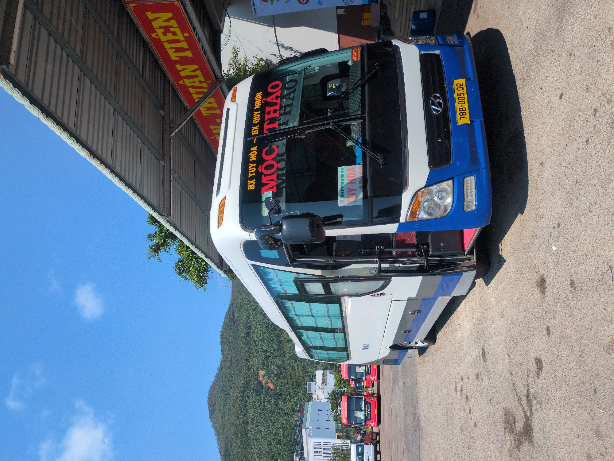 The image shows a white and blue Hyundai bus with the license plate 78B-005.02, parked on a paved surface. The bus has signage indicating it travels between Tuy Hoa and Quy Nhon, under the name "Moc Thao." In the background, there are more red buses and white buildings visible, with a green, tree-covered hill in the backdrop and a clear blue sky above. Part of a building structure with a sign saying "THUAN TIEN" is visible above the front of the bus. The image gives the impression of a bus depot or parking area. Based on the bus route and the company name, the location is South Quy Nhon Ward, Vietnam.