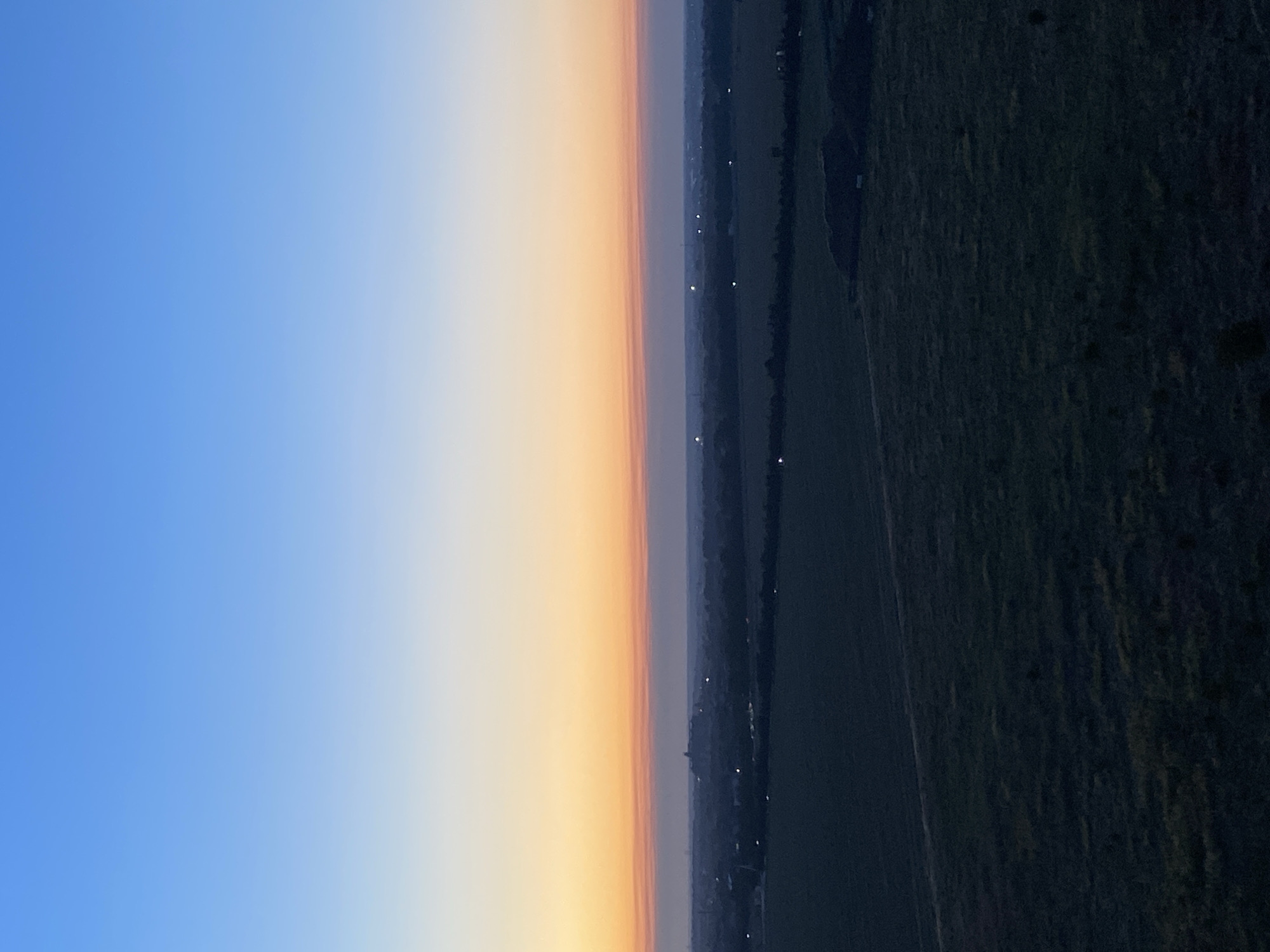 The image captures a twilight scene over a rural landscape. The sky transitions from a deep blue at the top to a soft orange and pink hue near the horizon, indicating either sunrise or sunset. Below the sky, a dark, silhouetted treeline separates the sky from the land. In the distance, faint lights suggest a settlement or town. The foreground is dominated by dark, textured fields, possibly farmland or open grassland, with some variations in shade and texture suggesting undulation or different types of vegetation. The overall impression is one of quietude and open space as day gives way to night, or vice versa. The image does not contain any identifiable people, animals, or specific structures that would allow for precise location identification within the city of Chornomorsk, Ukraine, based solely on visual elements.