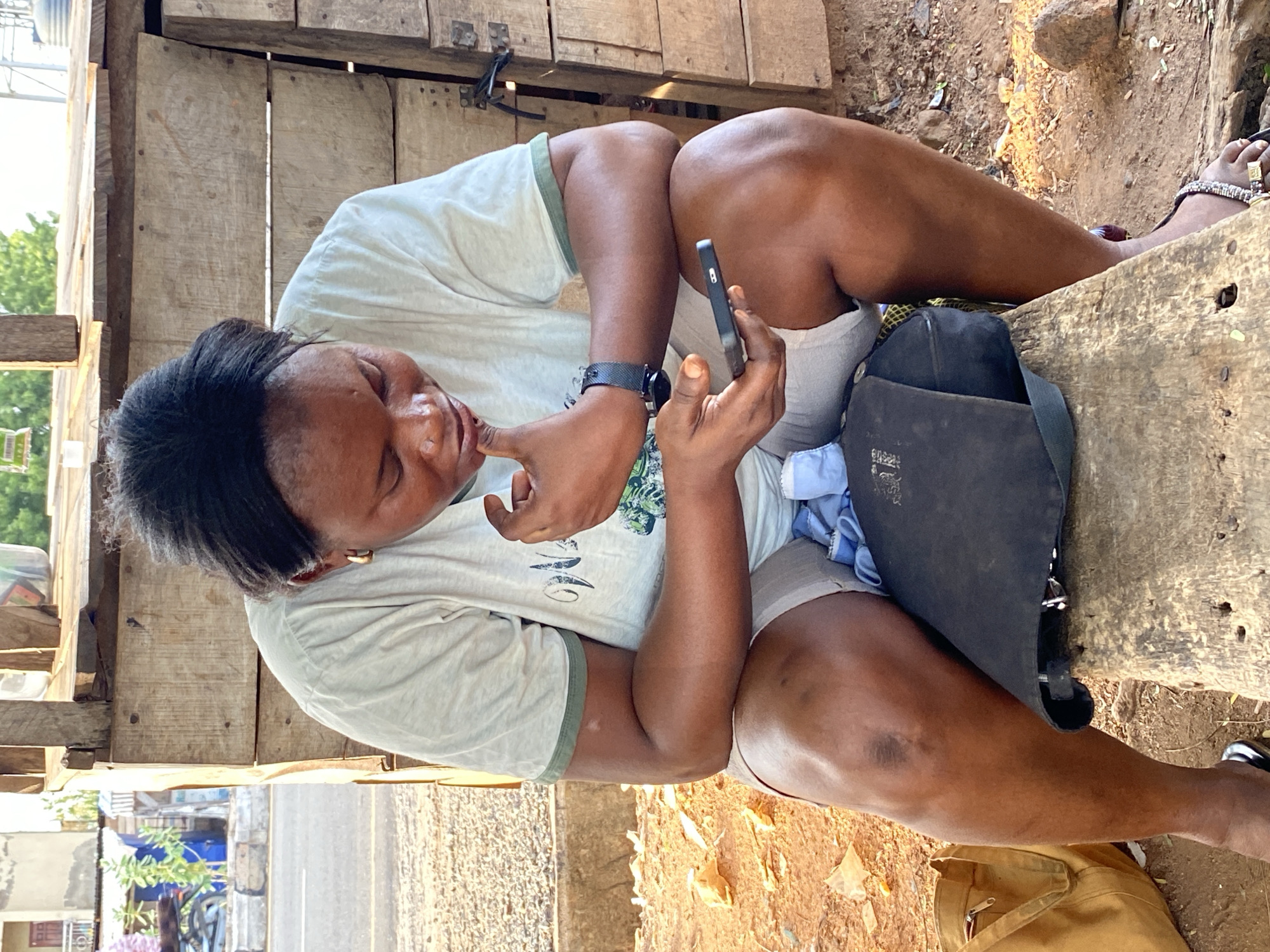 A woman of African descent is seated on a rustic wooden bench or platform, outdoors, under what appears to be a makeshift wooden shelter. She is dressed in a light-colored t-shirt with a faded graphic design and light-colored shorts or undergarments. She wears a dark wristwatch on her left wrist and sandals on her feet, with one foot adorned with beaded ankle jewelry.
The woman is actively engaged with a black smartphone, holding it in her right hand and looking intently at its screen. Her left index finger is poised near her mouth, suggesting concentration or contemplation. A black shoulder bag rests beside her on the wooden structure.
The setting appears to be a roadside or an informal marketplace in Adankolo, Nigeria, with dirt ground visible around the structure. In the background, there's a glimpse of a road or street, along with distant buildings and some greenery, suggesting a daytime, casual environment. The natural lighting indicates it is likely morning or afternoon.
The Apple logo is visible on the back of the smartphone. Faint, illegible text or symbols can be seen on the black shoulder bag.