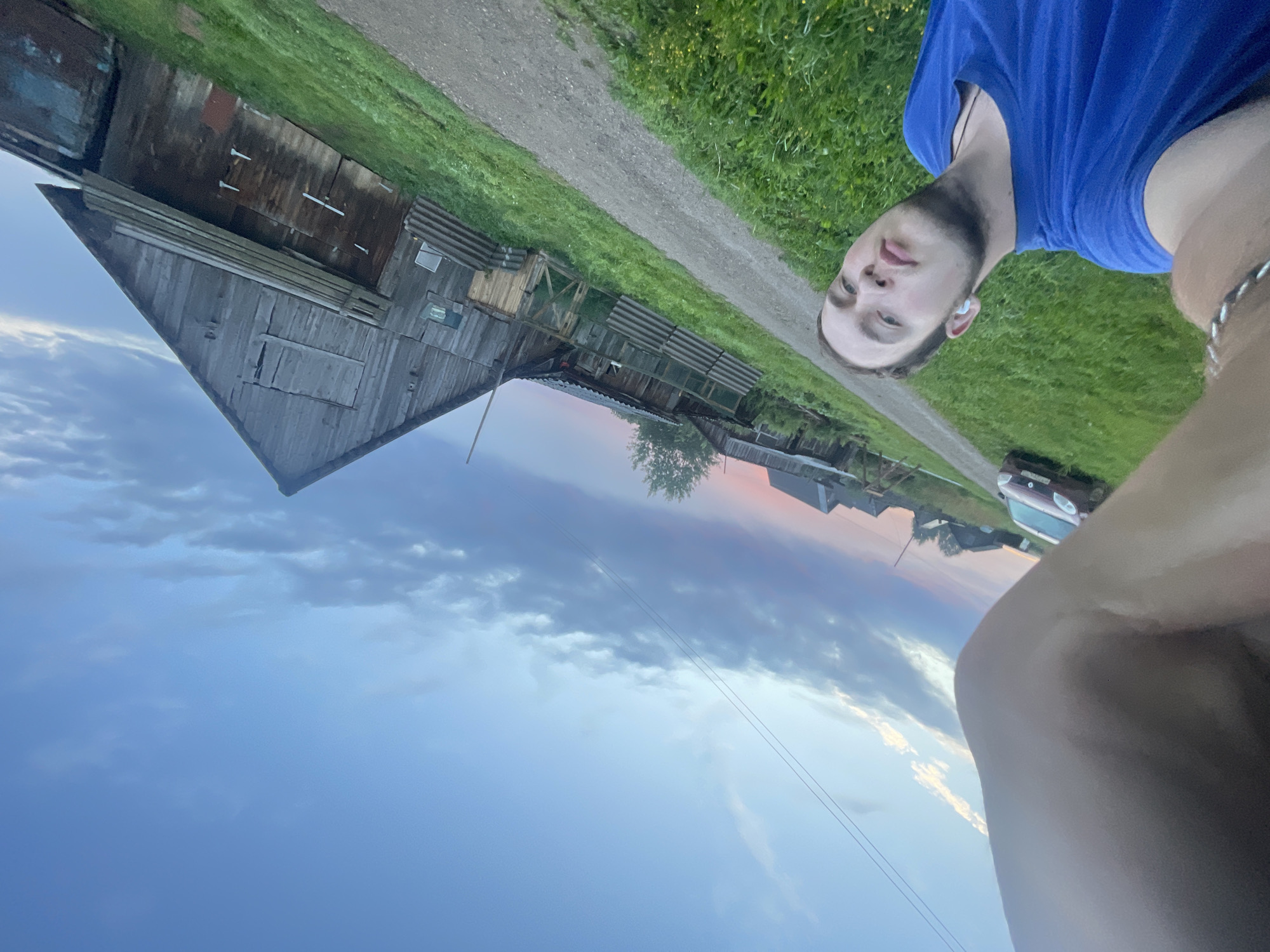 A young Caucasian male is taking a selfie with a rural Russian village in the background. He is wearing a blue t-shirt and earbuds. The village appears to be Panteleevka, Russia. The sky is a gradient of blue and pink, suggesting it is either sunrise or sunset. There are several wooden structures, including a barn, and a dirt road. A red car is parked on the left side of the road. The overall scene is one of a quiet, rural evening.