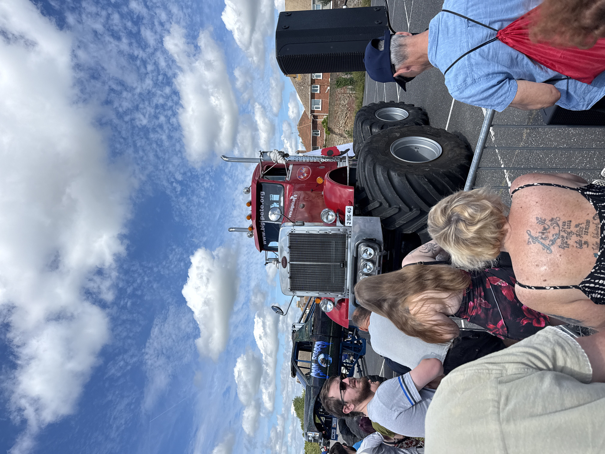 This media file captures an outdoor event, likely a truck show or community gathering, on a sunny day with a partly cloudy sky. The main subjects are a group of people viewing a large red and silver semi-truck with oversized tires. In the foreground, several individuals are visible from the chest up, with some looking towards the truck and others interacting amongst themselves. A woman with blonde hair and a floral dress is prominent, displaying a detailed tattoo on her arm and shoulder.
The truck, identified by visible text as "www.bigpete.org" and "2 BIG 4U," appears to be a centerpiece of the event. Another, more heavily modified truck with the words "ARIM REAPER" visible on its side, is partially seen behind the first truck. A large black speaker is positioned near the front of the scene. The setting appears to be a paved area, possibly a parking lot or street, with buildings in the background that suggest a suburban or town environment. The presence of a red drawstring bag worn by a man in a blue shirt indicates casual activity. The overall mood seems relaxed and celebratory, with people engaged in observing the vehicles and enjoying the outdoor atmosphere. The time of day is clearly daytime, and the weather appears to be pleasant for an outdoor event.