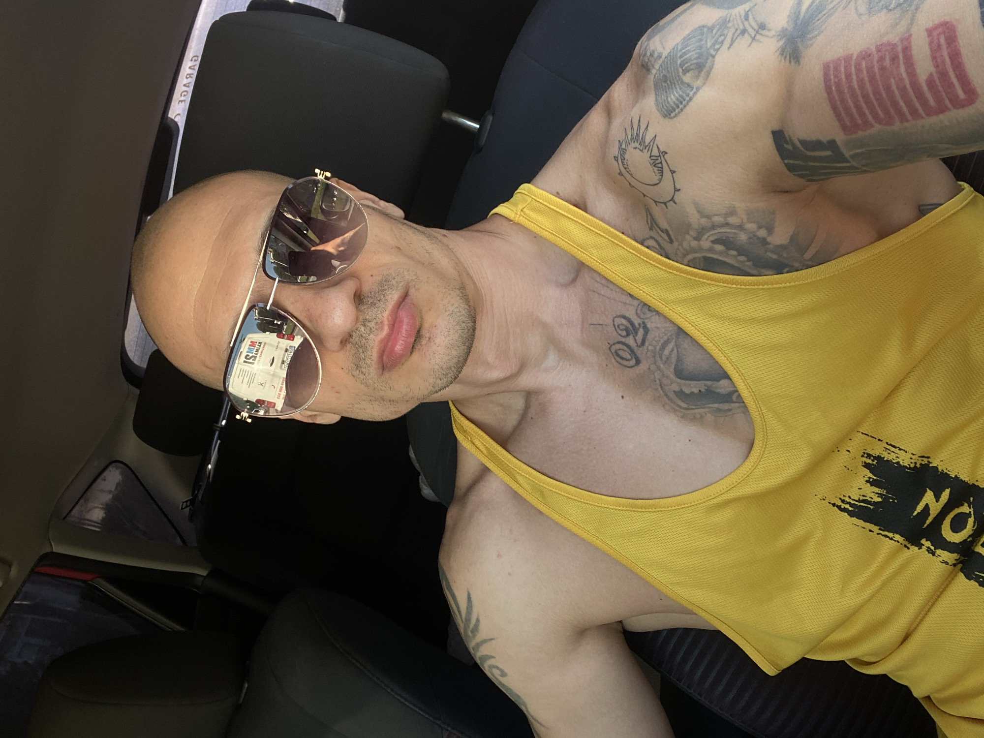 A man with a shaved head is in the front seat of a car. He is wearing sunglasses and a yellow tank top. The scene is inside a car with black interior. The reflection in his sunglasses indicates a commercial environment in Pleven, Bulgaria.