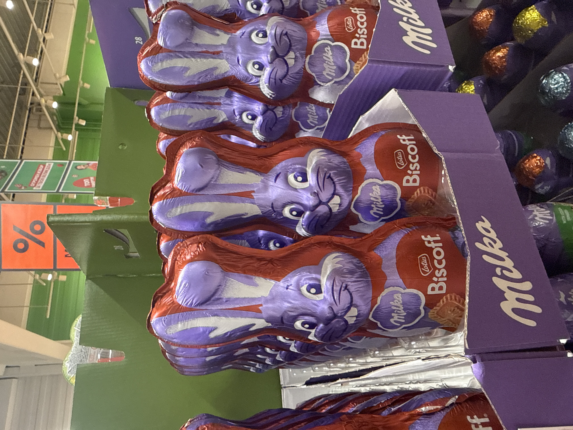 This is an eye-level, close-up shot of several boxes of Milka chocolate Easter bunnies wrapped in red and purple foil, stacked on a store shelf. The boxes are purple with the Milka and Lotus Biscoff logos. Behind the boxes is a green shelf and orange promotional signs with percentage symbols. In the background, there are more shelves stocked with foil-wrapped Easter eggs. The image appears to be taken indoors, likely in a supermarket. Given the presence of Cyrillic script on store signs, this is possibly taken in Bulgaria, but there is nothing indicating it is specifically Plovdiv.