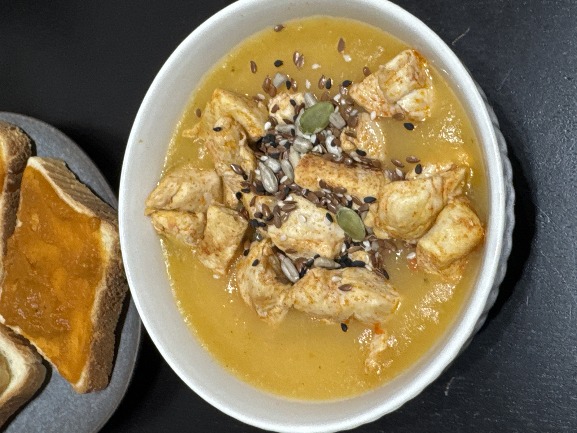 The image shows a bowl of yellow soup topped with chunks of cooked chicken and a sprinkle of mixed seeds (sunflower, pumpkin, flax, and black sesame). Next to the bowl, on a grey plate, are several slices of toasted bread, one of which is spread with an orange jam or marmalade. The background is a dark, possibly black, surface. There is no evidence in the image to suggest a location in Perm, Russia.