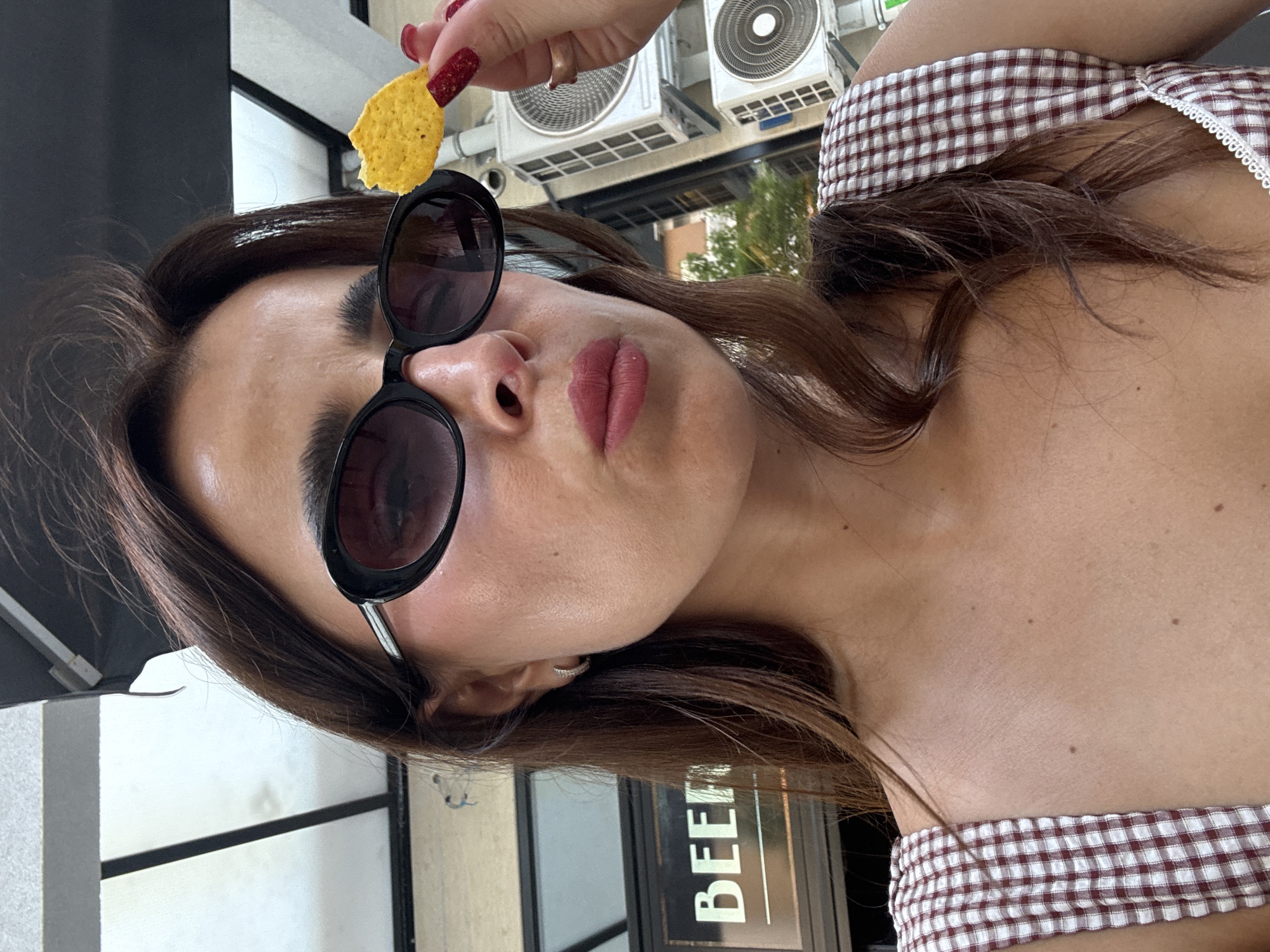 The image is a close-up, selfie-style shot of a woman wearing sunglasses and holding a single tortilla chip. She has long, dark brown hair and is wearing a red and white checkered top. Her lips are slightly pursed, and her fingernails are painted red. In the background, there is a sign that reads "BEEF" and what appear to be air conditioning units. The lighting suggests an outdoor daytime setting. The presence of the sign "BEEF" and the architectural style of the buildings in the background, including the visible air conditioning units, are consistent with an urban environment. While the image itself does not provide definitive evidence of the specific city of Podgorica, Montenegro, it depicts a scene commonly found in urban areas that could be located there.