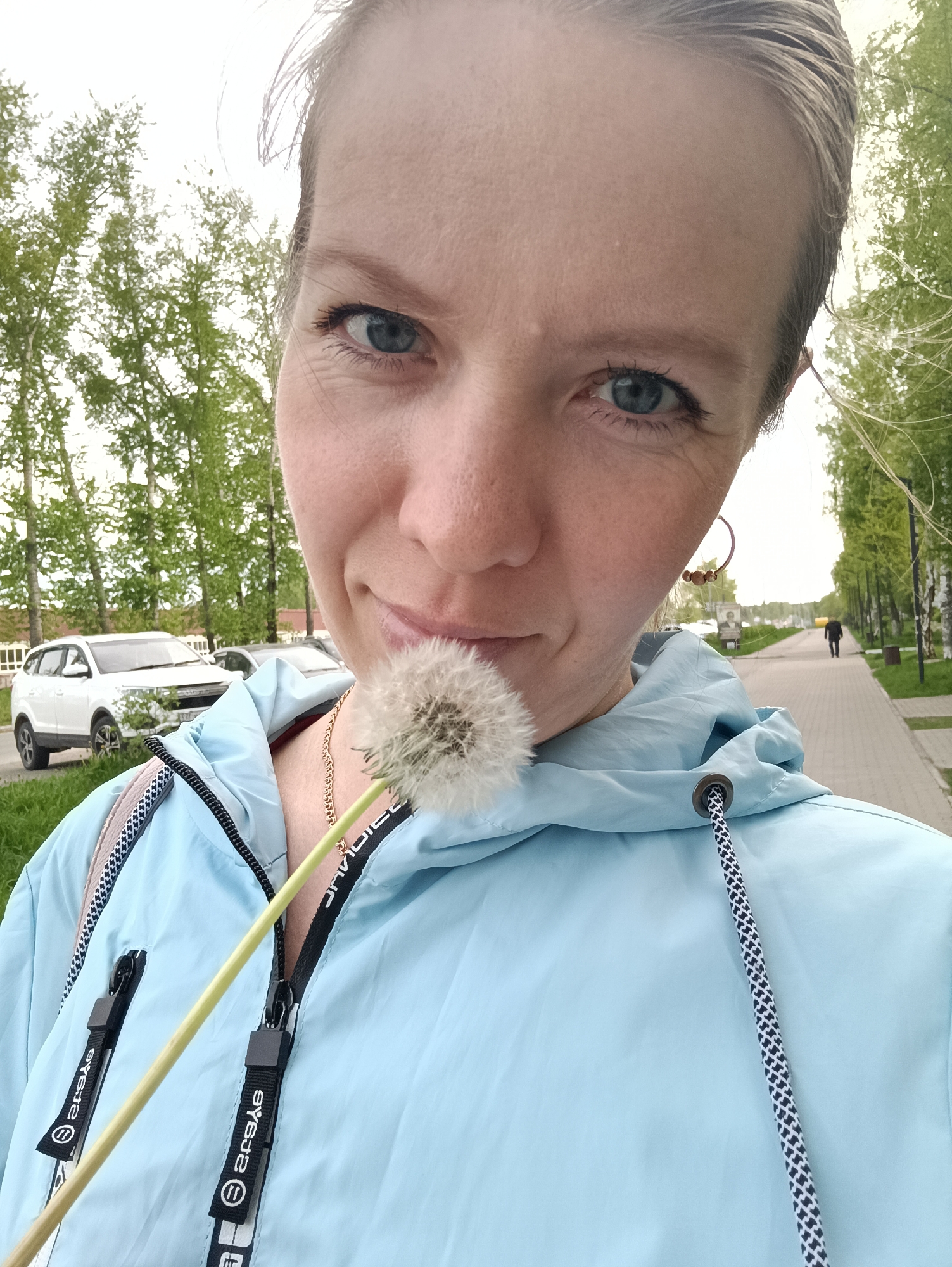 A woman with short blonde hair and blue eyes holds a dandelion in front of her face. She is wearing a light blue hooded jacket and a gold necklace. Behind her, a paved walkway stretches into the distance, lined with trees. A person walks on the walkway. To the left, a white car is parked near some green trees. The location is Chaikovsky, Russia.