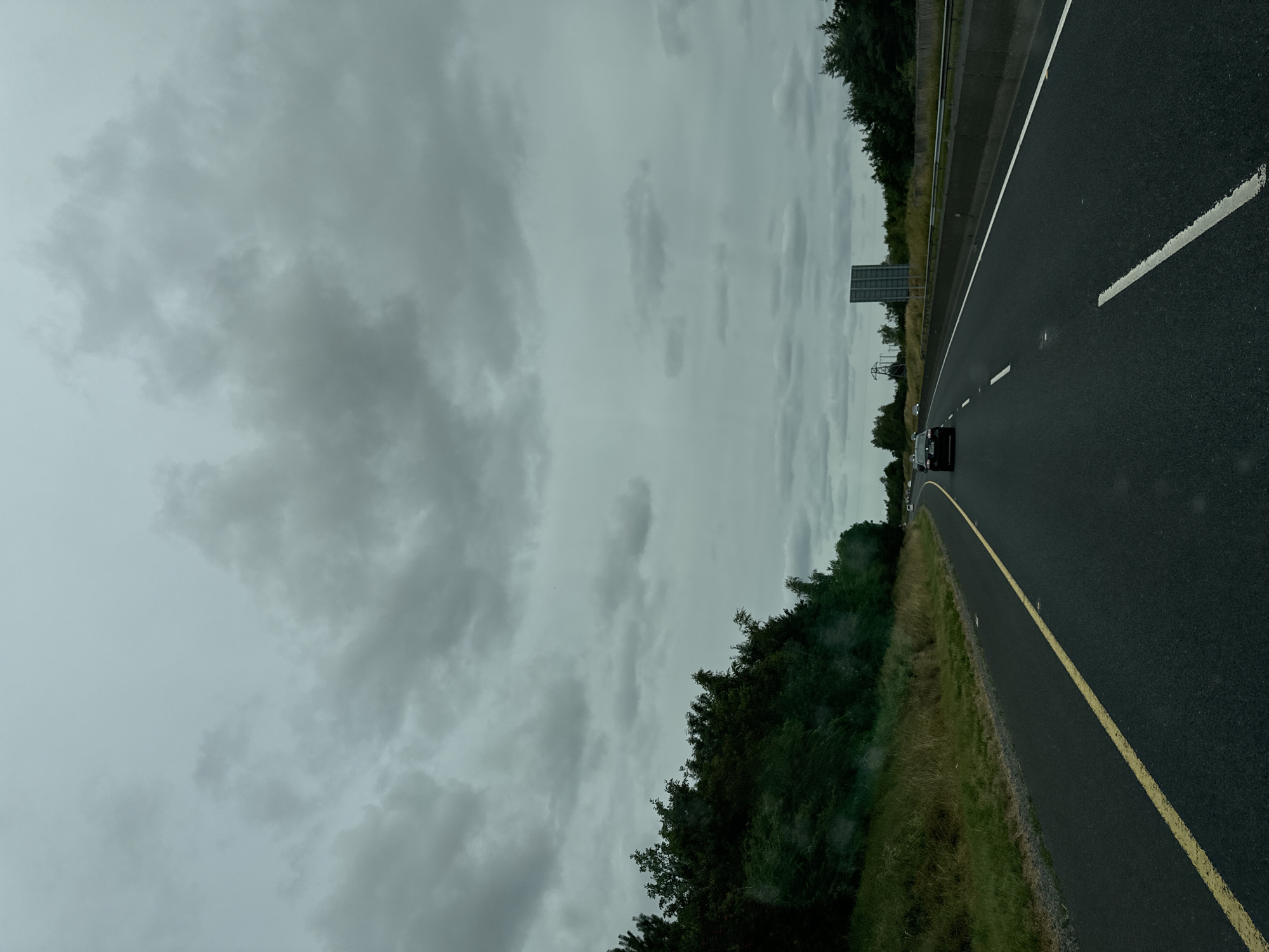 The image depicts a view from a vehicle traveling on a highway in the city of Old Kilcullen, Ireland. The sky is overcast with dark, heavy clouds. The highway is asphalt with clear lane markings, including a solid yellow line on the left and dashed white lines indicating lanes. A concrete barrier separates the highway from the opposite lanes of traffic. On the left side of the highway, there is a grassy embankment with lush green trees and vegetation. Several vehicles are visible on the highway, including a dark-colored car in the foreground and other cars further in the distance. A signpost is visible on the right side of the highway. Power lines and pylons are also visible in the background, suggesting the presence of infrastructure. The overall scene is one of a typical highway journey under a cloudy sky in a rural or semi-rural setting.