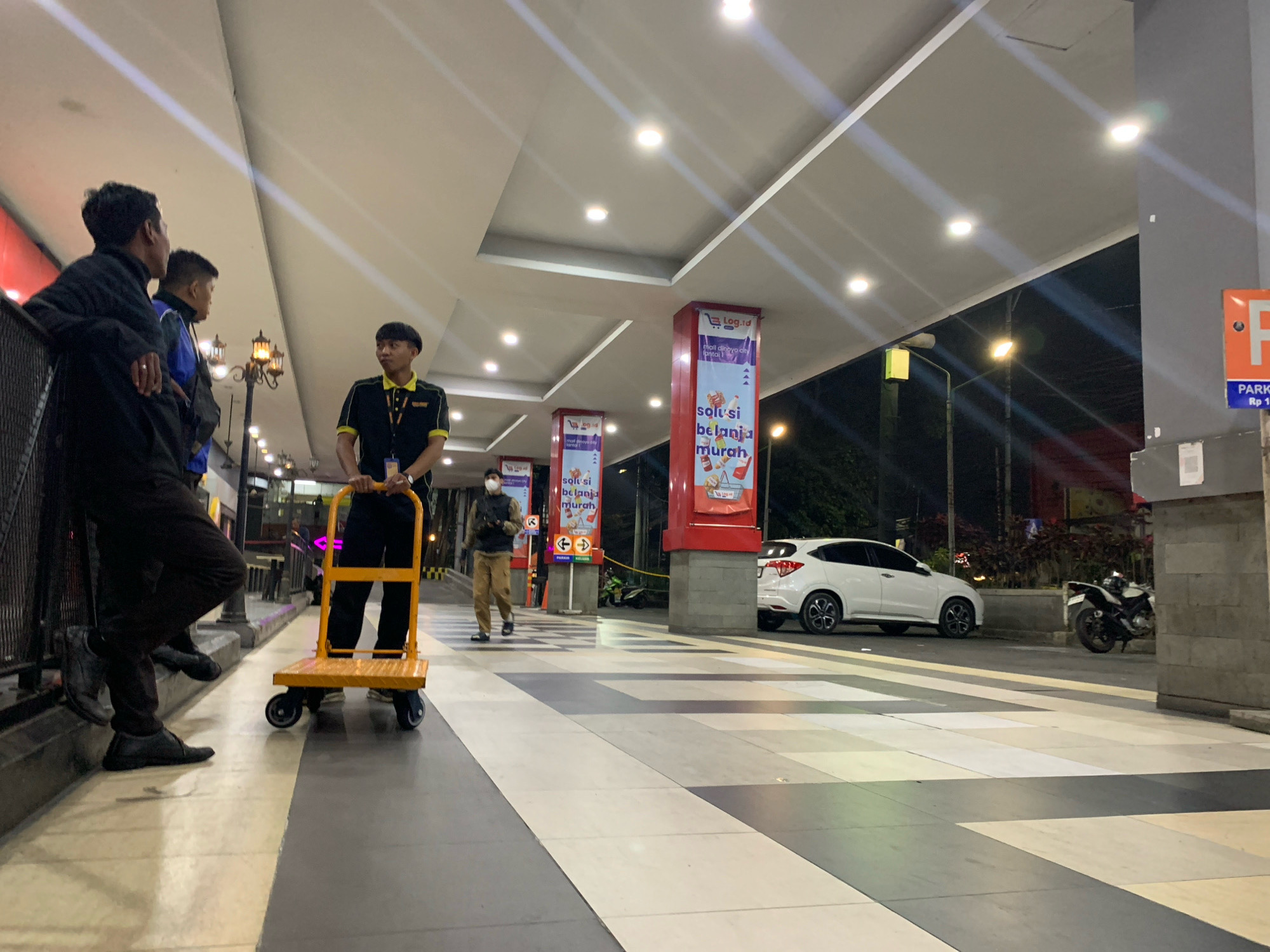 The image depicts an outdoor pedestrian walkway at night, likely in an urban setting in Malang, Indonesia. The walkway is paved with large, alternating light and dark gray tiles. Above, a white, modern canopy with recessed lighting provides illumination and casts diagonal light beams.
In the foreground left, three young men are standing. The man closest to the camera is wearing a black jacket, black pants, and black shoes, with his left leg crossed over his right. To his right, a second man in a blue jacket and black pants is partially visible. The third man, further back and to the right, wears a black shirt with a yellow collar and insignia, and dark pants. He is pushing a bright yellow, four-wheeled flatbed cart.
Further down the walkway, a man in a light brown uniform and dark pants walks away from the camera. He is wearing a face mask.
Red, vertical advertising pillars are spaced along the walkway, featuring graphics and text in Indonesian, including "Log.id" and "solusi belanja murah" (cheap shopping solutions). One pillar shows a shopping cart icon.
In the background, a white SUV is parked on a darker surface, possibly a road or parking area, adjacent to the walkway. To the right of the SUV, a black and white motorcycle is parked. Streetlights with orange-yellow bulbs are visible in the distance. A large, gray concrete pillar is on the far right, with an orange parking sign with the letter "P" in white, and blue text below indicating "PARKING Rp 1". The overall ambiance suggests a commercial or public area, possibly near a shopping center or mall.