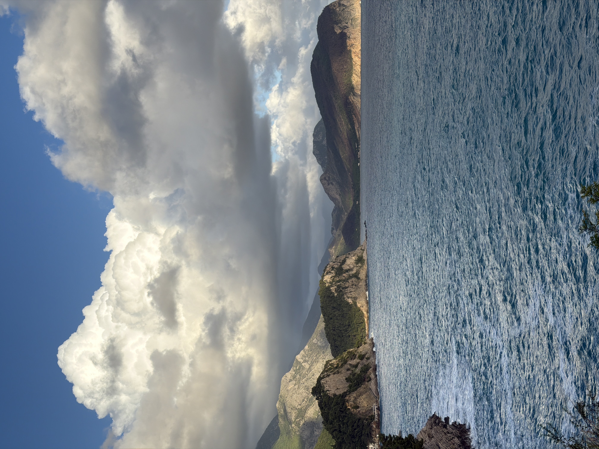 The image depicts a coastline in Mirište, Montenegro. A body of water, possibly the Adriatic Sea, occupies the right half of the frame, showing visible surface texture. The land is comprised of mountainous terrain with varying vegetation cover. A dramatic sky features billowing cumulus clouds on the left, contrasting with patches of blue sky. A column of rain falls from a dark cloud formation toward the center.