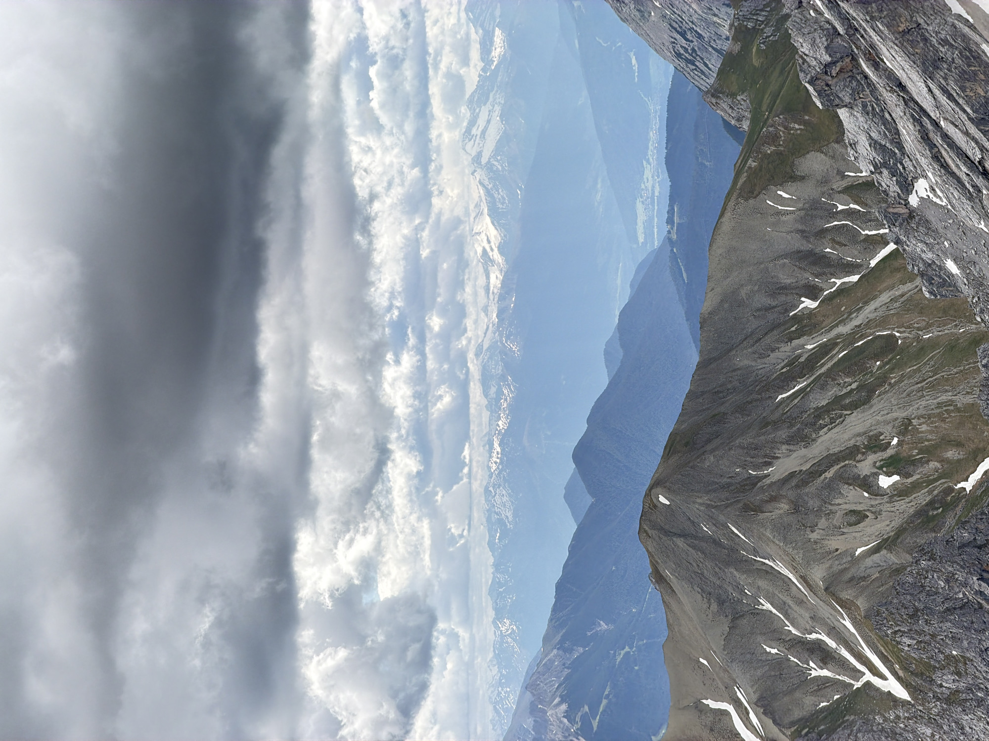 The image is a panoramic view of the Alps, likely from the Zugspitze mountain near Garmisch-Partenkirchen, Germany. The foreground features a rugged, rocky mountain slope with patches of snow and sparse green vegetation. In the middle ground, a series of dark blue mountain ranges recede into the distance, their peaks softened by atmospheric haze. The background is dominated by a vast expanse of sky filled with dramatic, dark gray storm clouds that partially obscure the distant, snow-capped peaks of the Alps. There are no people, animals, or man-made structures immediately visible in the foreground or middle ground, though a faint, distant village or town can be discerned in the valley between the mountain ranges. The lighting is subdued due to the overcast conditions, creating a somber and dramatic mood.
