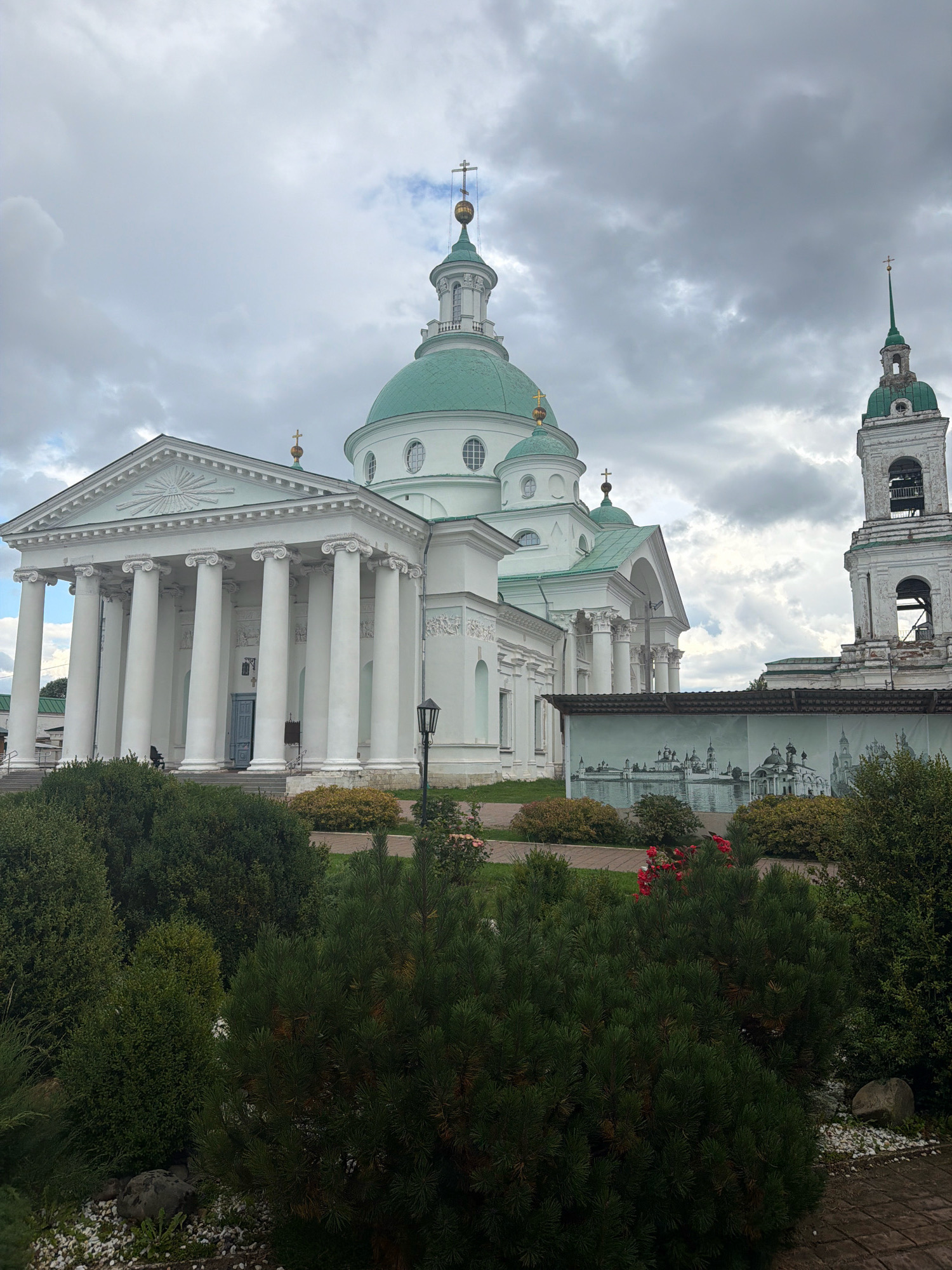 The image shows the Assumption Cathedral in the city of Kladovitsy, Russia. The cathedral features a large green dome with a golden cross at its apex. The facade of the cathedral is white with numerous columns and intricate architectural details. To the right of the cathedral, a tall bell tower with a green roof and a golden cross is visible. In the foreground, lush green bushes and trees are planted, with some red flowers adding a touch of color. The sky is overcast with gray clouds. A mural depicting various buildings and cityscapes is visible on a wall in the middle ground. A lamppost stands to the left of the mural.