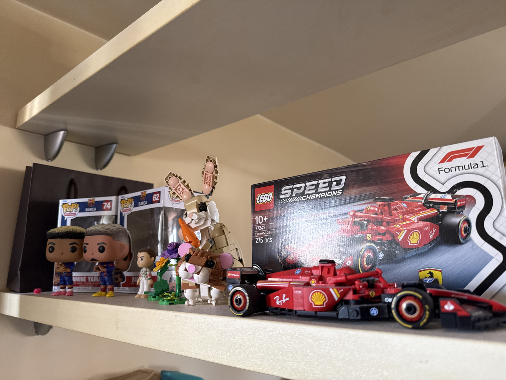 This indoor image, set in what is stated to be Sofia, Bulgaria, captures a collection of toys and collectibles displayed on a light-colored shelf against a warm beige wall. The perspective is low-angle, looking up at the shelf, which is supported by two sleek, silver-colored brackets.
On the left side of the shelf, a dark bag is partially visible behind two Funko Pop! figures. These figures depict male football players in blue and red striped uniforms, consistent with FC Barcelona. One figure, numbered 74, is associated with "AMINE YAMA" (likely Lamine Yamal), while the other is numbered 62 with "BARÇA" visible on its box. A small pink object rests on the shelf nearby. To their right stands a smaller figure in a white patterned shirt.
The central part of the display features Lego creations. A large, intricate Lego rabbit, built in shades of brown and tan, holds an orange carrot. Smaller Lego plant elements are arranged around its base. In front of the rabbit is a smaller, brown Lego creature.
The right side of the shelf is dominated by a Lego Speed Champions set. A prominently displayed box for the "LEGO SPEED CHAMPIONS" "Ferrari SF-24" (set 77242, 275 pcs, 10+) shows an image of the racing car, along with the "Formula 1" logo. In front of the box, the assembled red Lego Ferrari SF-24 car is showcased. This model features sponsor decals including "V-Power," "Ray-Ban," and a "Shell" logo, matching details on the box.
The image is a static portrayal of a personal collection, with no discernible activity or specific emotions, relying on natural or indirect indoor lighting.