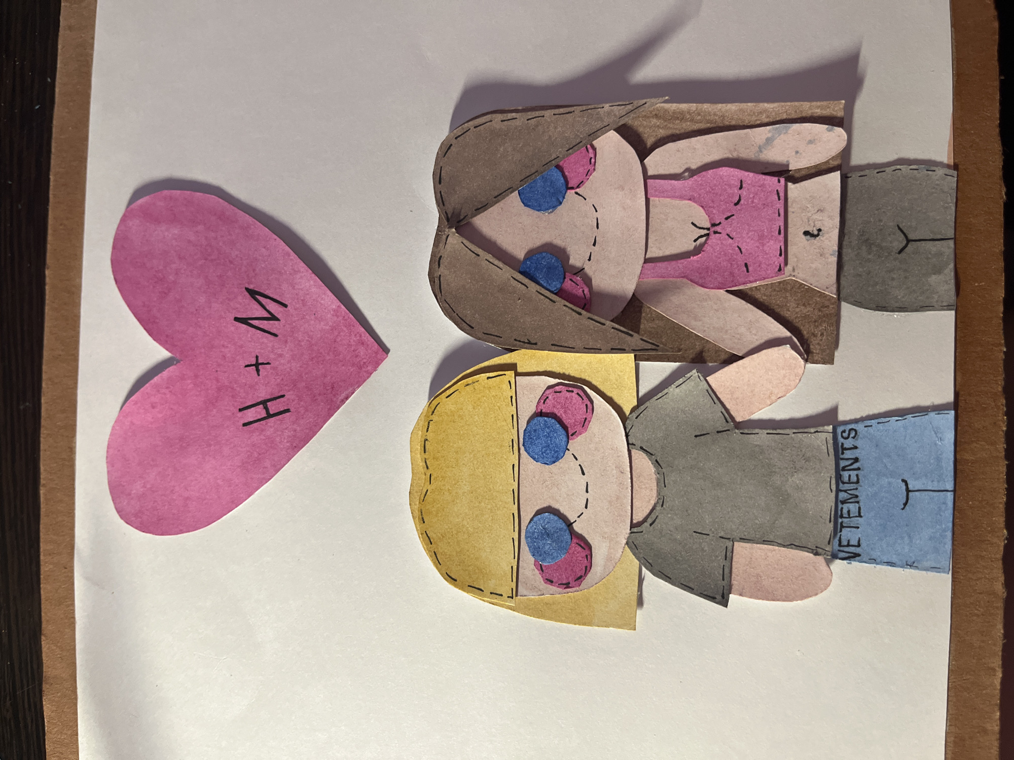 This image depicts a handmade collage on a white background, possibly a card or a piece of artwork. The collage consists of two stylized figures and a pink heart.
The figures appear to represent a couple, constructed from cut paper or cardstock. One figure has brown hair, a pink top, and grey pants. The other has blonde hair, a grey top, and blue pants with the word "VETEMENTS" written on it. Both figures have pink cheeks and blue eyes. The "VETEMENTS" is written vertically.
To the left of the figures is a pink heart shape. The letters "H + M" are written on the heart.
There is no evidence to suggest the location is in Saint Petersburg, Russia.