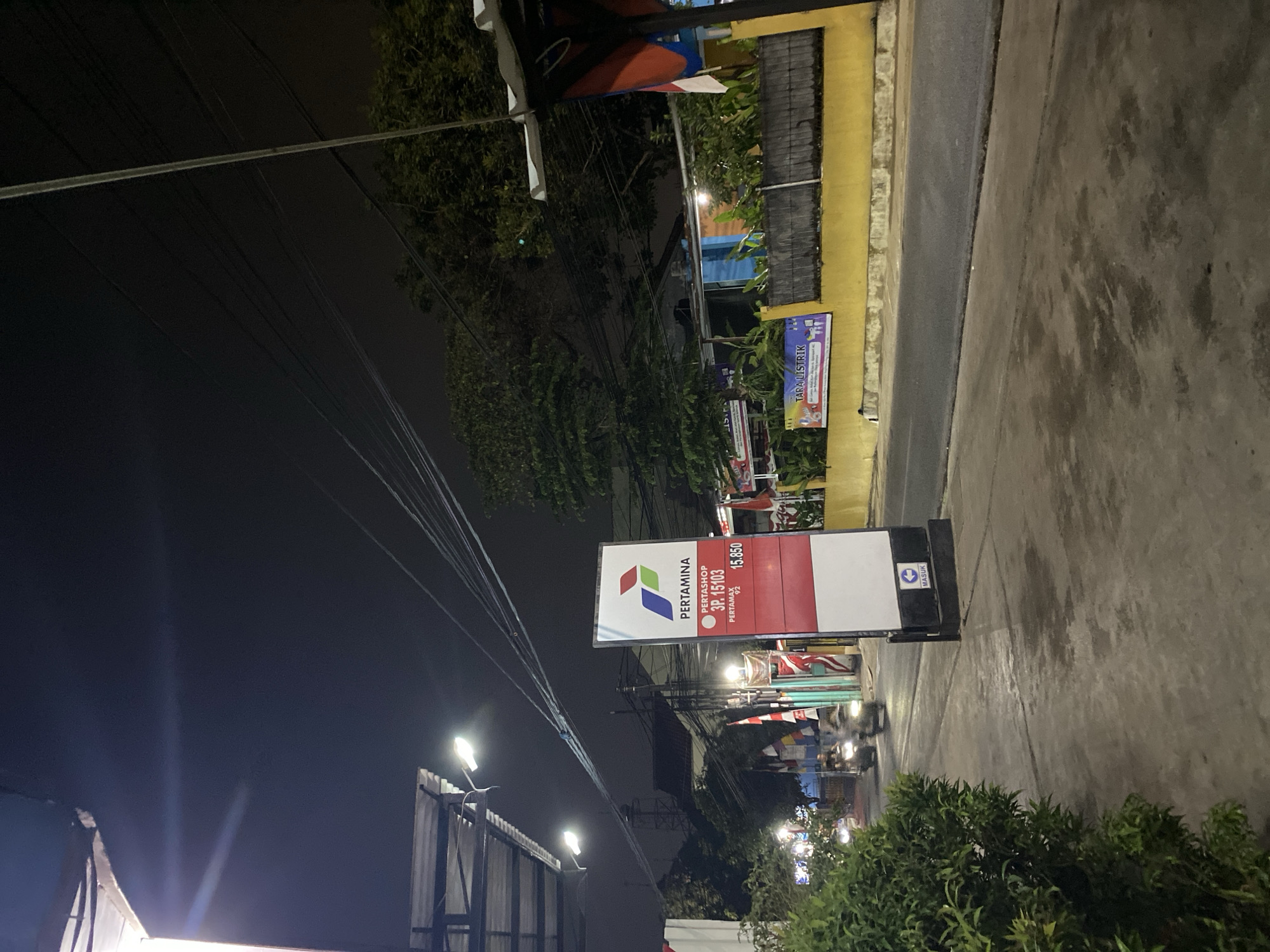 This is a nighttime photograph of a Pertamina gas station sign on a street in Tangerang, Indonesia. The sign is tall and rectangular, with a white background and the Pertamina logo at the top. Below the logo, it lists "PERTAMAX", "SP. 15103", and "15.850" which likely refers to the price per liter. The sign also displays a "MASUK" (entrance) sign with an arrow pointing left.
To the left of the sign, a sidewalk is visible with a green bush in the foreground. A street runs parallel to the sidewalk, with a yellow wall and a wooden fence in the background. Several Indonesian flags are displayed on poles along the street. The sky is dark with a few lights visible, and power lines crisscross the scene overhead. Two motorcycles are moving on the street, blurred due to motion. Buildings and trees are visible in the background. The overall impression is of a well-lit gas station entrance at night.