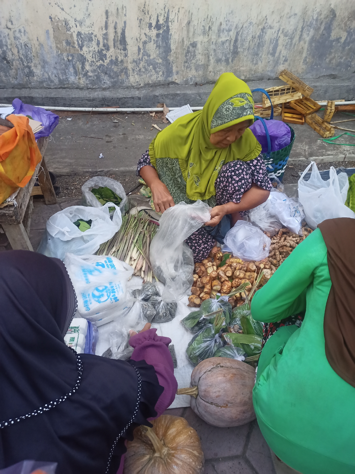 In Nganti, Indonesia, a market scene unfolds with multiple individuals, likely vendors, engaged in commerce. A woman in a lime green hijab and floral-patterned dress is seated on the ground, surrounded by various produce. She is in the process of handling a plastic bag filled with goods. To her right, another person, their face obscured by a brown hijab and wearing a green garment, is also present. In the foreground, a third individual, wearing a black hijab and a purple sleeve, is visible. Several pumpkins, a variety of roots and spices, and bunches of lemongrass are displayed on the ground. Bags of green leafy vegetables and what appear to be smaller, darker produce are also present. Wooden crates are stacked behind the seated woman, and plastic bags are scattered around. The ground is paved with interlocking bricks, and a concrete wall forms the backdrop.