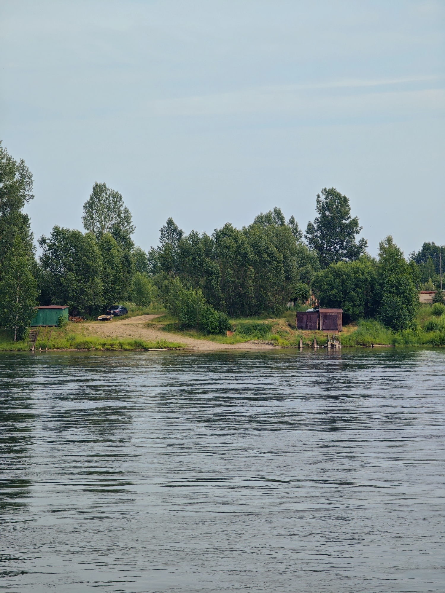 The image depicts a serene river scene with a lush green shoreline under a pale, overcast sky. On the bank, several trees with dense foliage dominate the landscape. Near the water's edge, a small, green, shed-like structure stands on stilts, with wooden steps leading up to it. Further along the bank, a dark-colored car is parked on a sandy area, next to a small trailer. Two rusty, metal-clad structures, resembling storage sheds or small garages, are situated closer to the water, with wooden steps descending towards the river. The river itself is calm, with gentle ripples on its surface, reflecting the muted light and the greenery of the opposite bank. The overall impression is one of a quiet, somewhat rustic, riverside location. This scene could potentially be located in the vicinity of the city of Sosnovoborsk, Russia, which is known for its proximity to the Yenisei River and its natural landscapes.