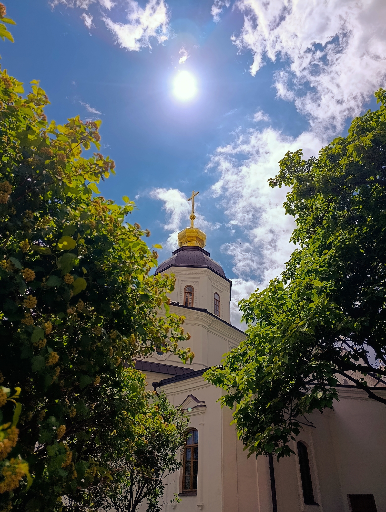 The image shows the exterior of a church with a golden dome and cross, framed by lush green trees. The sky is blue with white clouds, and the sun is shining brightly, creating a halo effect. The architecture of the church is visible, with white walls and arched windows. The trees are full of leaves, and some have small yellow flowers. The overall impression is one of a serene and beautiful day. The church is the Kyiv Pechersk Lavra, a historic Orthodox Christian monastery located in Kyiv, Ukraine.