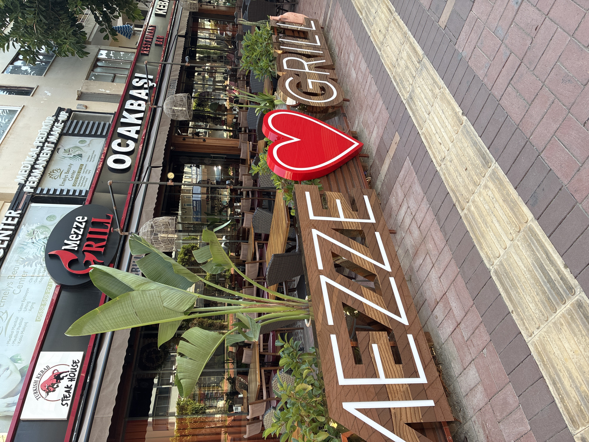 In Alanya, Turkey, a storefront displays the name "MEZZE GRILL" in large wooden letters with white outlines, positioned at street level. A prominent red heart sculpture separates the word "MEZZE" from "GRILL." Behind these letters, a restaurant with outdoor seating is visible, featuring tables and chairs. Overhead, a striped awning covers the dining area. Signs above the awning advertise "OCAKBAŞI RESTAURANT BAR" and "KEBAB." Further up, a sign for "Emelys Beauty Center" and "MEDİKAL PEDİKÜR PERMANENT MAKE-UP" is visible. A tall, leafy plant stands in front of the Mezze Grill signage. The foreground shows a sidewalk paved with interlocking bricks of various colors, including a section with tactile paving for visually impaired pedestrians. The scene is captured in daylight, suggesting an outdoor urban setting.