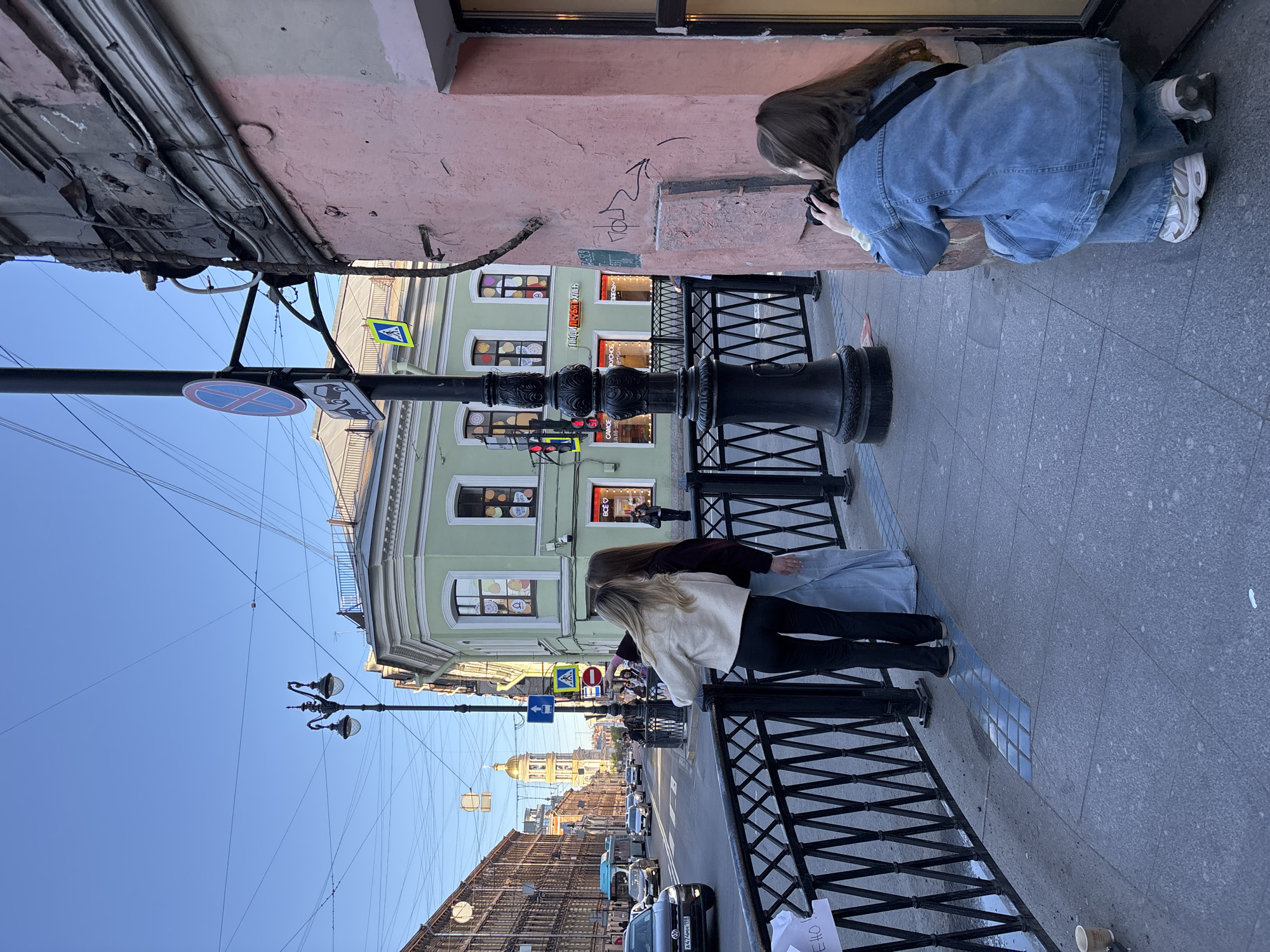 Two women are on the sidewalk of a street in Saint Petersburg, Russia. One woman is crouched, taking a photo of graffiti on a pink wall. The other is leaning against a black metal fence, looking down the street. The street is lined with buildings, and cars are driving along it. A gold-domed building can be seen in the distance. The sky is blue, and several street signs are visible on poles.