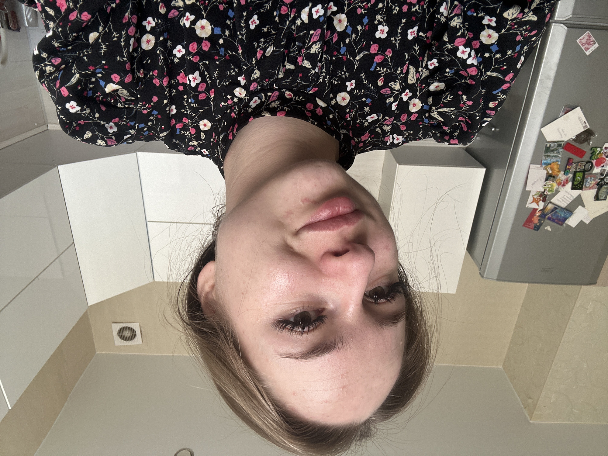 A young adult female is depicted in a close-up, high-angle selfie photograph taken indoors in what appears to be a residential kitchen in Unknown, Unknown. The subject has light brown hair, dark eyes with prominent dark eyelashes, a fair complexion with minor blemishes on the forehead and around the mouth, and appears to be smiling faintly. She is wearing a black long-sleeved top with a high mock neck, adorned with a vibrant floral pattern consisting of pink, white, and light blue blossoms with green foliage. The top features subtly puffed shoulders. To the left of the subject, a silver-grey two-door refrigerator, bearing the 'beko' brand name, is visible. Its upper door is covered with numerous colorful decorative magnets and one small white envelope. The background consists of light beige walls with a subtle textured pattern, meeting a white ceiling. White upper kitchen cabinets with a modern design are visible along the right side of the frame. A square white wall-mounted ventilation fan with a circular grid is positioned on the wall in the upper right corner of the room. The overall lighting is bright and even.