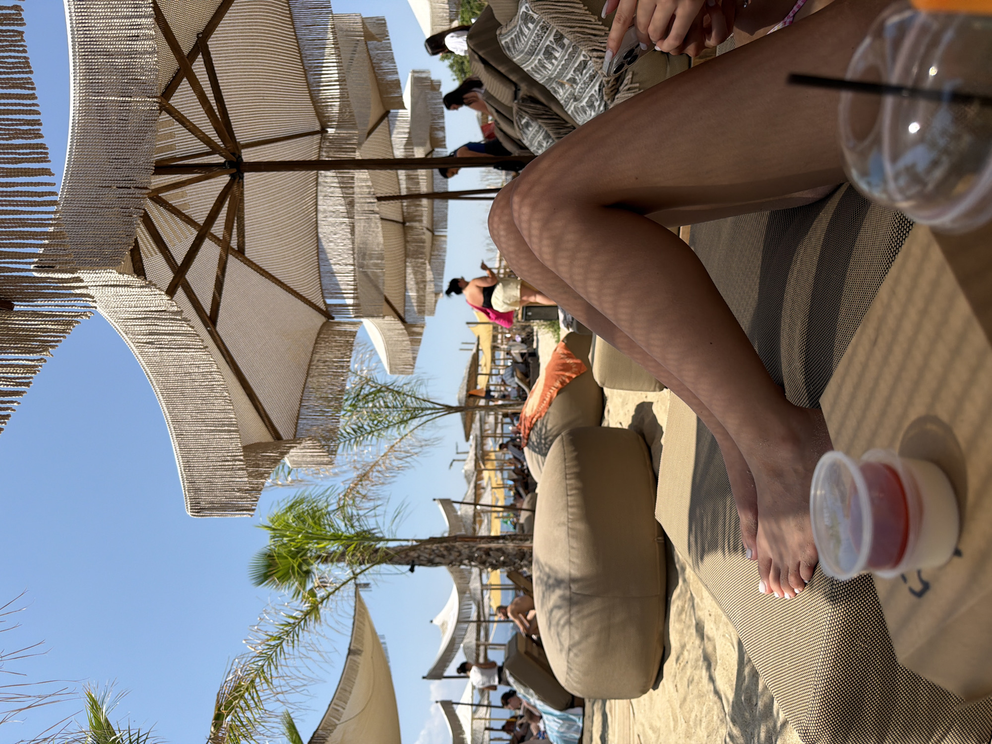 The image depicts a sunny day at Ofrynio Beach, Greece. The foreground is dominated by a person's legs, lying on a sun lounger. A small plastic container with a red dessert and a clear plastic cup with a straw are on a surface in front of the lounger, casting shadows from an overhead umbrella. In the mid-ground, beach chairs and large, circular, tan beanbag chairs are scattered across the sand. Several people are visible in the background, some sitting on loungers and others standing. Palm trees and more umbrellas, featuring fringed edges, are scattered throughout the beach scene. The sky is a clear, bright blue.