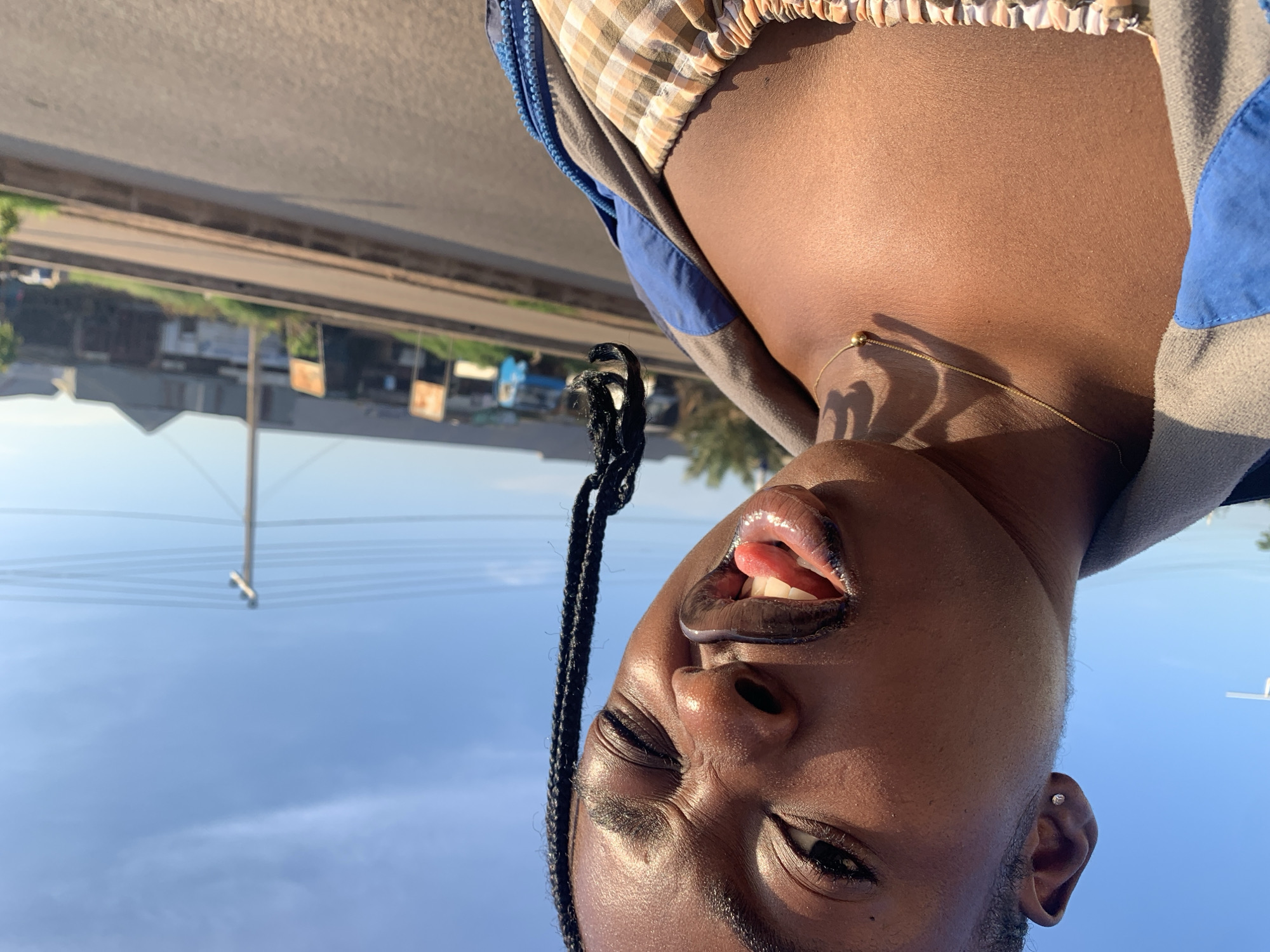 A close-up, selfie-style portrait features a young Black person with braided hair, sticking their tongue out and winking, set against a bright blue sky with sparse clouds. The person wears a grey and blue jacket zipped over a tan and white plaid garment. A delicate gold chain with a single bead rests on their collarbone. The background, slightly out of focus, depicts a street scene with a paved road, a curb, and distant buildings and structures, possibly indicating an urban or semi-urban environment in Rafin Par, Nigeria. A utility pole with multiple wires is visible on the right. The lighting suggests it is daytime, with sunlight highlighting the person's skin.