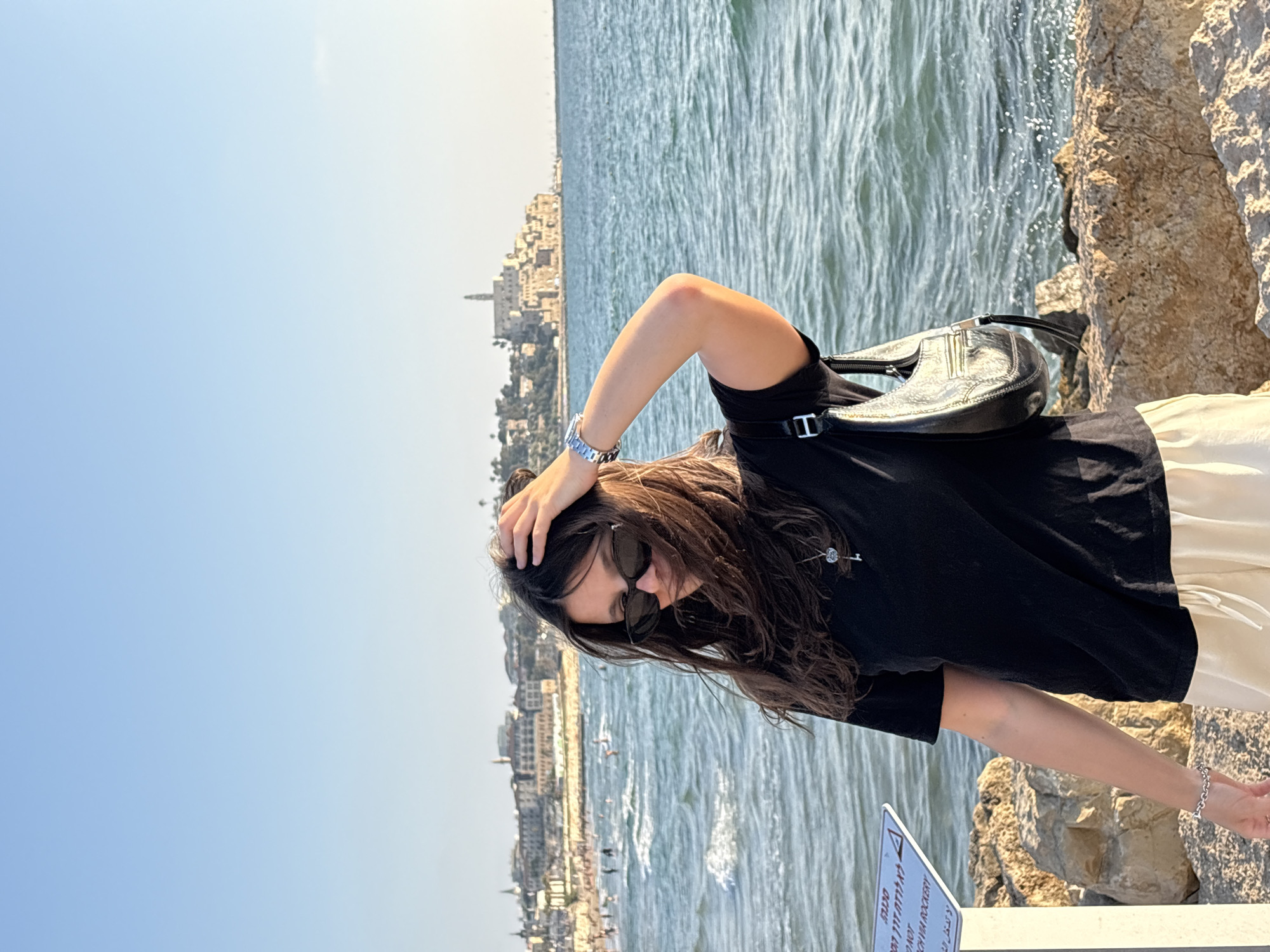 A young woman with long, dark hair and sunglasses stands on a rocky shore, looking down and adjusting her hair with her right hand. She wears a black t-shirt, light-colored shorts, and a silver bracelet on her left wrist. A black shoulder bag hangs from her right shoulder. Behind her, the blue sea is visible with small waves, and in the distance, a city skyline with buildings and a prominent tower is seen. The sky is clear and blue. A sign with text in Hebrew and English is partially visible on the left side of the image. The scene appears to be Jaffa Port in Tel-Aviv, Israel.