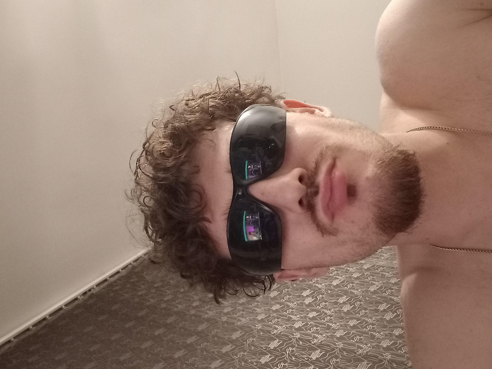 The image shows a close-up, selfie-style shot of a young man wearing dark sunglasses. He has curly brown hair and a goatee. His expression is neutral, with his lips slightly pursed. The sunglasses reflect a screen displaying what appears to be a video game or online content with multiple windows. The background is a wall with patterned wallpaper on the left and a plain, light-colored wall on the right, meeting at a corner. A thin chain necklace is visible around his neck. The image appears to be taken indoors. There is no visible indication of the city of Parvomay, Bulgaria.