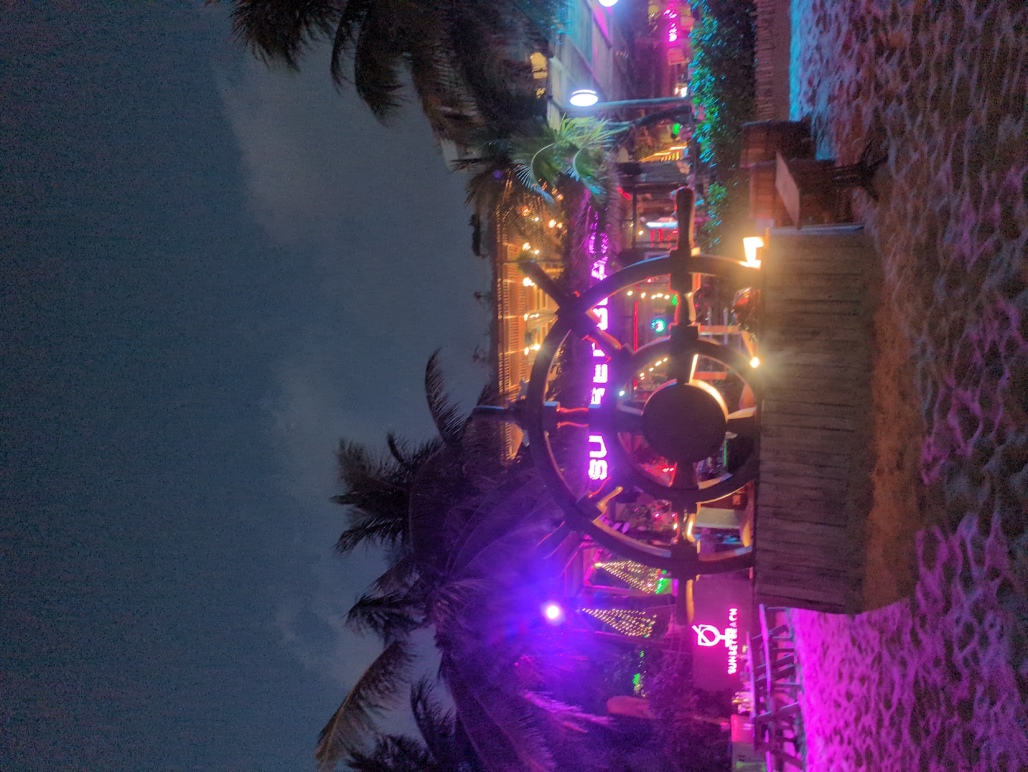 The image depicts a scene at the Sunset Beach Bar in Phú Quốc, Vietnam, at night. A large wooden ship's wheel, illuminated with purple neon lighting, is a central feature. Palm trees are visible on either side of the wheel, silhouetted against the night sky. Behind the wheel, the bar area is illuminated with various neon lights. The ground is covered in sand and is illuminated in pink light. The sky above is dark with hints of clouds.
