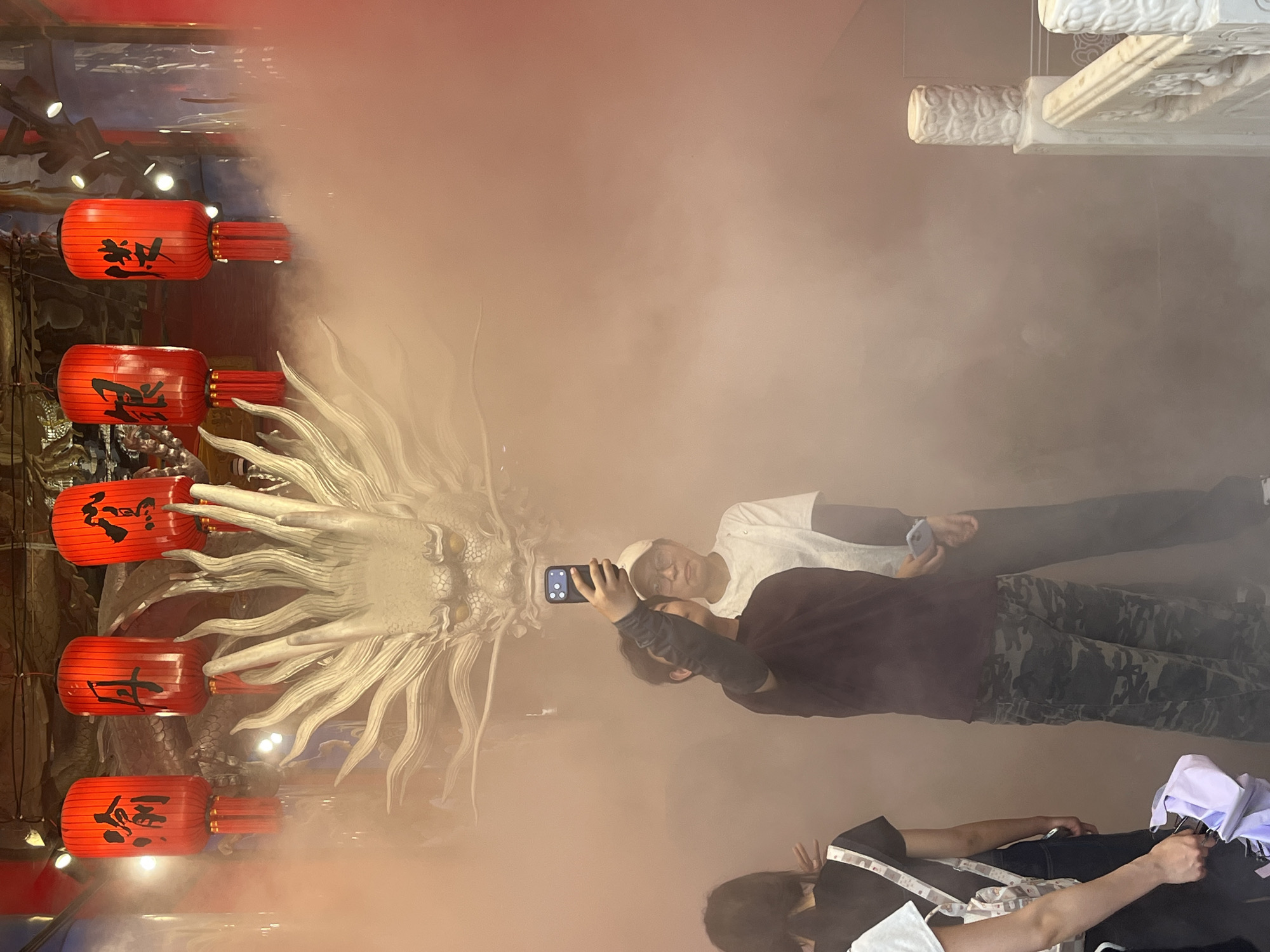 In the Yuzhong District of China, a group of individuals stands before a large, ornate dragon head sculpture. The sculpture appears to be a centerpiece of the scene, with smoke or fog gently swirling around it, creating an ethereal atmosphere. Hanging above the dragon are several traditional red Chinese lanterns, each adorned with black calligraphy.
In the foreground, three people are visible. One person, facing away from the camera and holding a white bag, is on the far left. To their right, a person in a dark shirt and camouflage pants is taking a selfie with their phone. Behind them, another person wearing a white t-shirt and a white baseball cap is looking towards the camera. The overall setting suggests an indoor cultural or decorative space, possibly a temple or a themed attraction, characterized by its traditional Chinese elements and dramatic lighting and atmospheric effects.