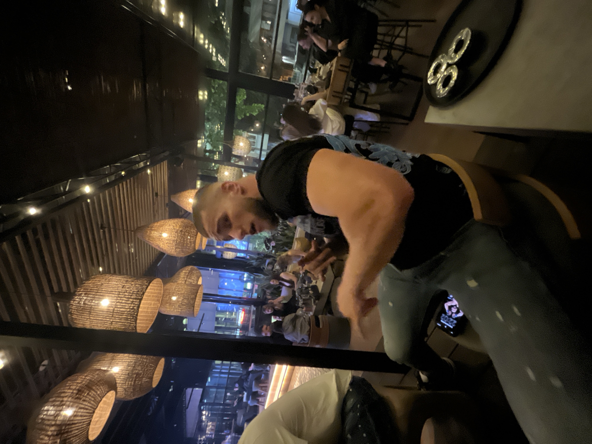 The image shows a man in a restaurant setting. The location is identified by the presence of external surroundings similar to the city of Burgas, Bulgaria.
The man is wearing a black t-shirt and blue jeans. He is partially in the foreground, with the restaurant extending behind him. The restaurant appears to be dimly lit, with decorative lighting fixtures. Other individuals are present in the background, seated at tables. There's also a tray on a table in the upper right corner.