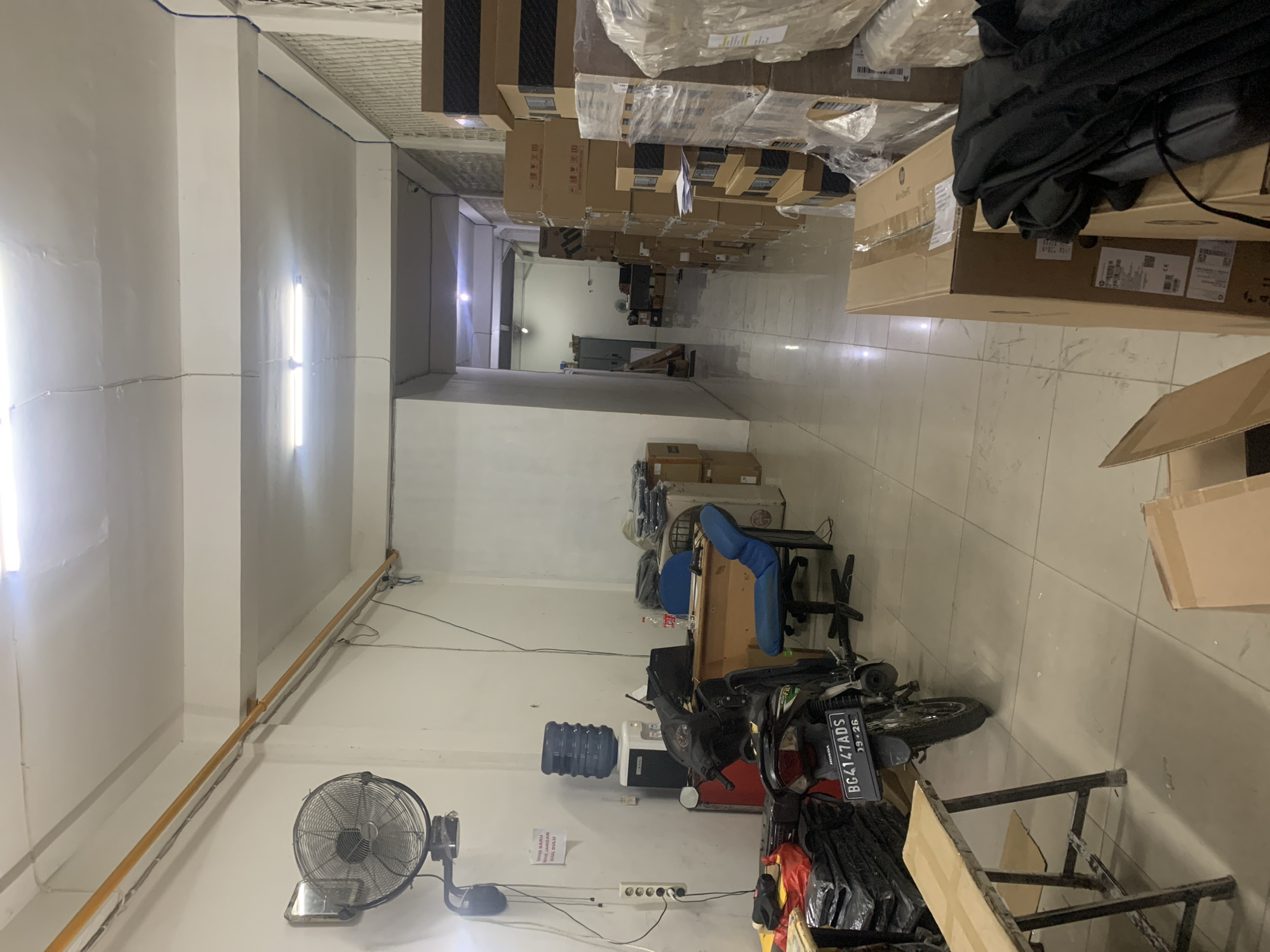 The image depicts the interior of a small warehouse or storage room, likely in Palembang, Indonesia. The space is narrow and long, with a white tiled floor and white walls. Fluorescent lights are visible on the ceiling. On the left side of the room, a wall-mounted fan is prominent, above a water cooler with a large blue water jug. A black motorcycle with a license plate reading "BG 4147 ADS" is parked in the foreground. Stacked black items are visible on a wooden table. Further back, a wooden desk with blue office chairs and a laptop is present. The right side of the room is lined with numerous cardboard boxes of various sizes, some wrapped in plastic. These boxes are stacked high, suggesting storage of goods. The overall impression is one of a functional, albeit somewhat cluttered, storage facility.