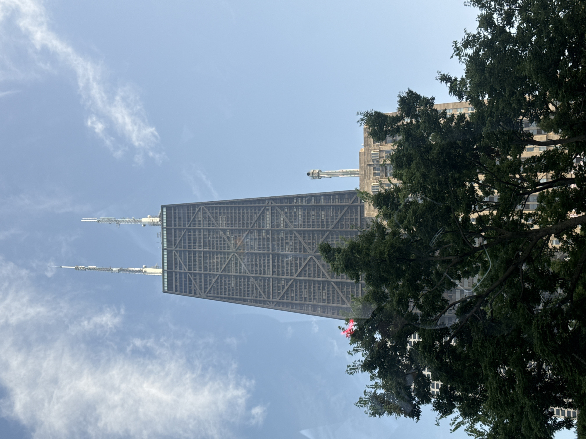 The image captures a low-angle view of the John Hancock Center in Chicago, Illinois, United States of America. The iconic skyscraper, known for its distinctive cross-braced exoskeleton, dominates the frame, reaching towards a clear blue sky with wisps of white clouds. Two prominent antenna towers stand atop the building, extending further into the sky. In the foreground, the lush green foliage of trees partially obscures the lower sections of the skyscraper, creating a natural contrast with the urban architecture. The lighting suggests it is daytime, with the sun illuminating the scene from an unseen angle. The overall impression is a characteristic Chicago urban landscape, where towering architecture meets natural elements.