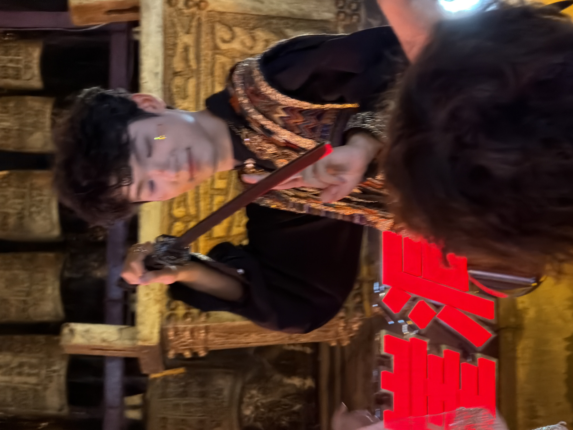The image shows a young man holding a sword in what appears to be a performance or ceremonial setting. He has dark, curly hair and is wearing a black outfit with an ornate, gold-embroidered vest. There are large, bell-like objects in the background, possibly part of a traditional musical instrument or decoration. Large red Chinese characters are visible in the foreground, partially obscuring the lower portion of the scene. The overall lighting suggests an indoor or evening event. The location is identified as Yuzhong District, China.