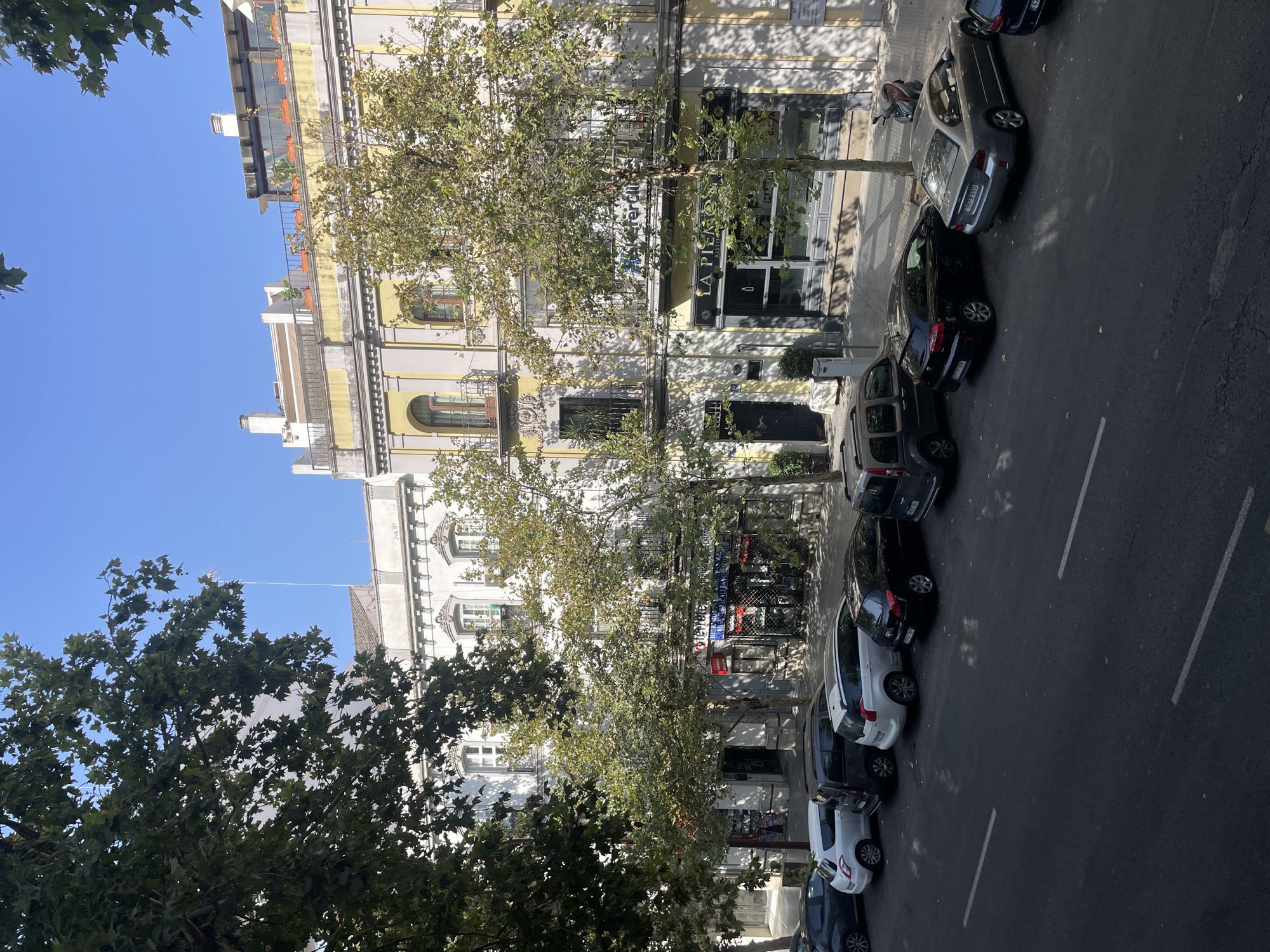 The image shows a street scene in Seville, Spain, on a sunny day. In the foreground, a street is lined with parked cars, including a white hatchback, a dark grey minivan, a white station wagon, a black hatchback, and a silver sedan. In the background, there are several buildings with traditional architecture, featuring balconies, arched windows, and ornate detailing. Trees with green foliage line the street, casting shadows on the pavement. The sky is clear and blue. A scooter is parked on the sidewalk. The building in the center has a sign that reads "LA PIERRO RISTORANTE."