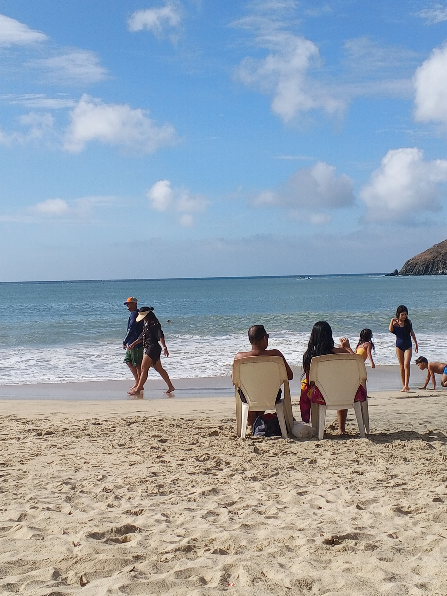 The image captures a beach scene in Pedro González, Venezuela. The scene is composed of a sandy beach, the ocean, a partly cloudy blue sky, and a land formation in the distance to the right.
In the foreground, a man and a woman are sitting on white plastic chairs. Behind the chairs and slightly to the right, two young girls and a boy are seen near the water. One girl is standing, while the boy is crawling near the water. To the left, a man and a woman are walking along the water's edge. The man is wearing an orange hat and a blue shirt, while the woman is wearing black clothing and a sun visor. The water is receding onto the beach in small waves. The sand is tan and slightly damp. The sky is blue with scattered white clouds. In the distance, a land formation appears to be covered in vegetation.