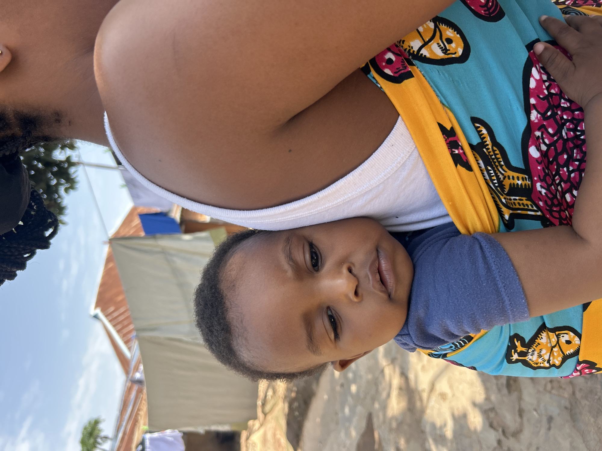An extreme close-up shot focuses on a young Black child being carried on their mother's back. The child, with dark curly hair, is leaning against the mother's shoulder and has a thoughtful expression. They are wearing a blue t-shirt and are wrapped in a colorful fabric sling with patterns of yellow, turquoise, and pink. The mother's right shoulder and upper arm are visible, wearing a white tank top. The background suggests a modest outdoor setting, possibly in a village or informal settlement, with a clothesline holding laundry, corrugated metal roofs of buildings, and dusty ground. The lighting indicates it is daytime with a bright, clear sky. The focus is tight, with the child and sling occupying most of the frame, blurring the background details. The image does not provide enough specific visual information to definitively pinpoint the location within Jos, Nigeria, but the general scene is consistent with rural or semi-urban areas in West Africa.