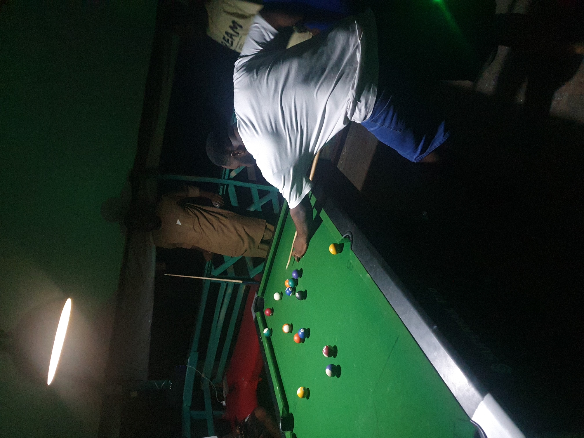 In Lokoja, Nigeria, a group of individuals are gathered around a green billiards table in a dimly lit indoor setting, likely a recreational facility or a private establishment. A prominent fluorescent light fixture illuminates the scene from above. The primary focus is on a man wearing a white t-shirt and blue denim shorts, actively engaged in playing billiards. He is positioned at the table, holding a cue stick and preparing to strike a billiard ball. Several other billiard balls of various colors are scattered across the green felt surface. In the background, two other individuals are visible. One man is wearing a light-colored, possibly tan, traditional robe, and appears to be observing the game. Another person is partially visible on the right, wearing a light-colored t-shirt with the word "TEAM" printed on it. The environment suggests an informal gathering, possibly after dark, with limited artificial lighting creating distinct shadows. The background shows a glimpse of what appears to be a green-painted structure, possibly a fence or seating area, and a red-colored floor or mat. The table itself has markings, including "SUPERMAX PRO" embossed along its side. The overall atmosphere is relaxed and social, centered around the activity of playing billiards.