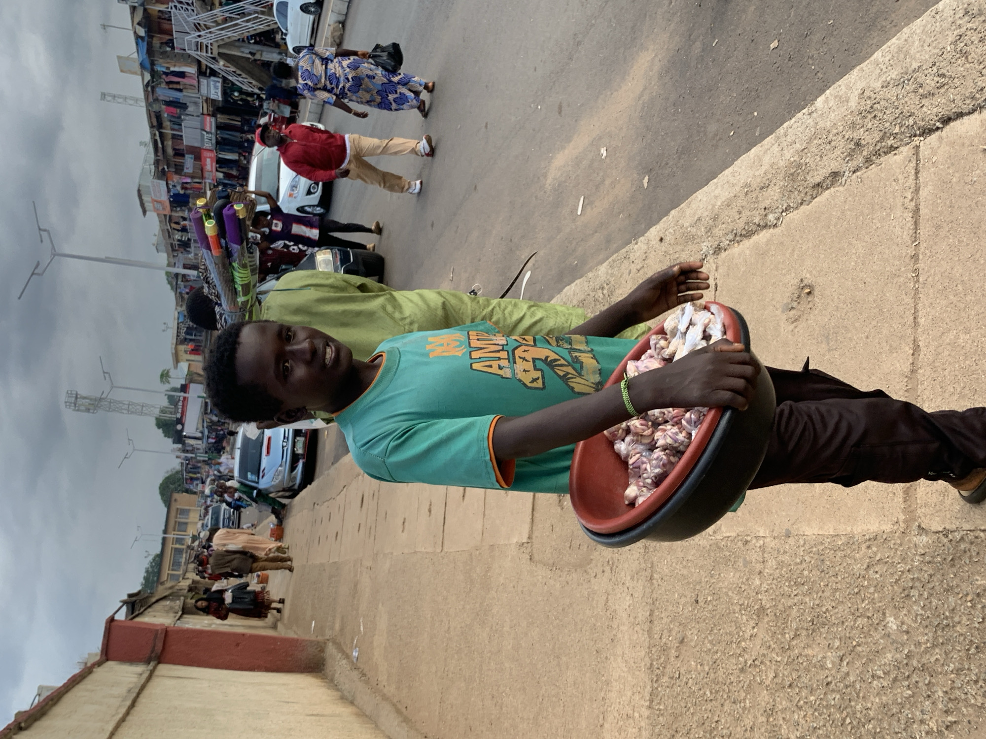 In Jos, Nigeria, a young boy stands on a sidewalk, holding a red basin filled with raw meat. He wears a green t-shirt with orange trim and dark pants. Behind him, a bustling market scene unfolds with stalls lining the street, displaying clothing and goods. People walk along the sidewalk and a street with vehicles, including a silver car and a white car. The sky is overcast.