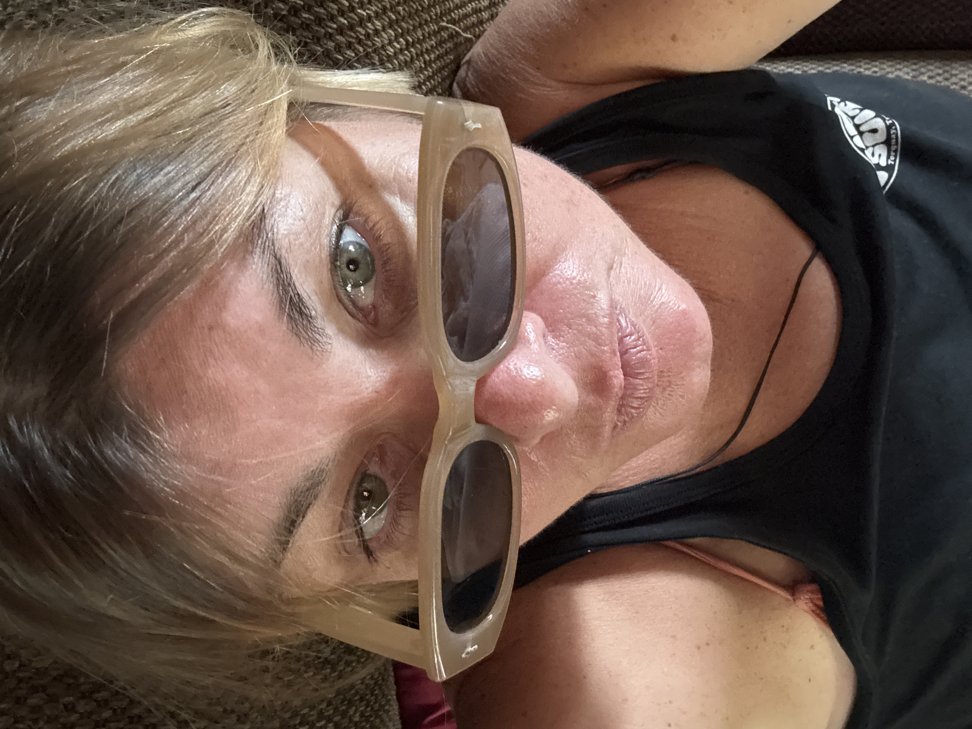 A close-up, overhead shot features a woman with blonde hair and sunglasses resting on her nose. Her green eyes are visible, looking upwards. She is wearing a black tank top. The background appears to be a textured brown surface, possibly a couch or chair. The lighting suggests an indoor setting.