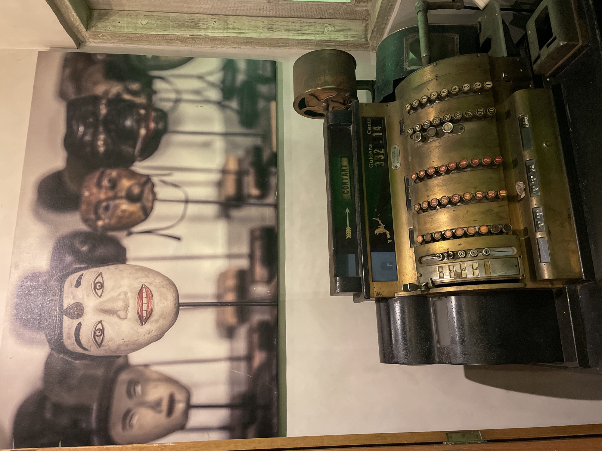 This indoor shot shows a vintage cash register mounted on a wall next to a framed photograph of traditional Indonesian masks. The cash register is made of brass and dark metal, with buttons and levers still intact. It displays "Guldens Cents 332.14" on its display. Next to it is a framed photo of a collection of Indonesian masks with distinct painted features. The location is in Kuta Selatan, Indonesia.