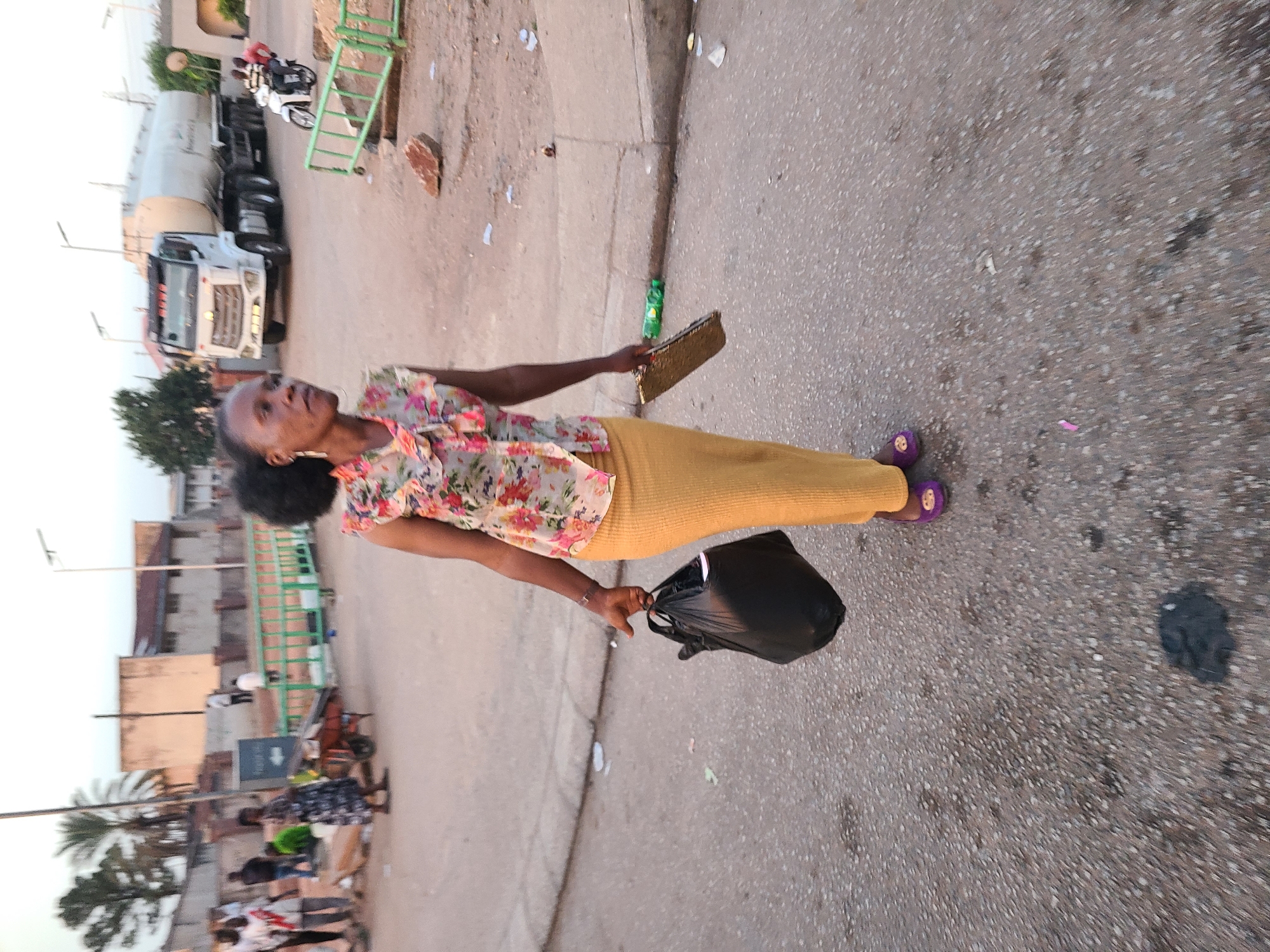 A woman is walking on a street in Unknown, Unknown. She is wearing a floral top, a yellow skirt, and purple sandals. She is carrying a black plastic bag in her left hand and a gold clutch in her right hand. The street is littered with trash. In the background, there is a truck and some buildings.