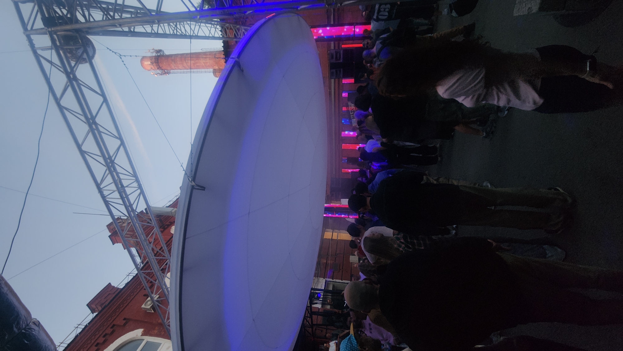 The image depicts an outdoor evening event or gathering at an industrial complex in the city of Unknown, Unknown. The scene is illuminated by artificial light against a twilight sky.
A large, circular white fabric canopy is suspended overhead, glowing with a prominent purplish-blue hue, suggesting direct illumination from below. This canopy is supported by a visible metal truss system.
In the background, multi-story red brick buildings with an industrial aesthetic are visible. On the left, a building features arched windows and an external air conditioning unit. On the right, a tall, slender red brick industrial chimney with an external ladder or scaffolding extends towards the sky, further reinforcing the industrial setting. Additional metal support structures and cables span across the upper portion of the frame.
Beneath the canopy, a crowd of numerous individuals is gathered, standing on what appears to be a dark paved surface. Most people are viewed from behind or from a slight side angle, appearing as silhouettes or dimly lit figures. They wear casual attire, including t-shirts, hoodies, and caps. No specific actions or interactions beyond standing and observing are clearly discernible among the crowd.
Further back, beyond the immediate crowd and beneath the canopy, a performance or display area is visible. This area features multiple tall, vertical rectangular light panels emitting bright red and pink/magenta light. A banner with "HAZE" is faintly visible above these panels. Speaker enclosures are also present on stands to either side of this lighted section.
The overall lighting is low, with the artificial lights of the event providing the primary illumination and casting a distinct glow on the overhead canopy and the background structures.