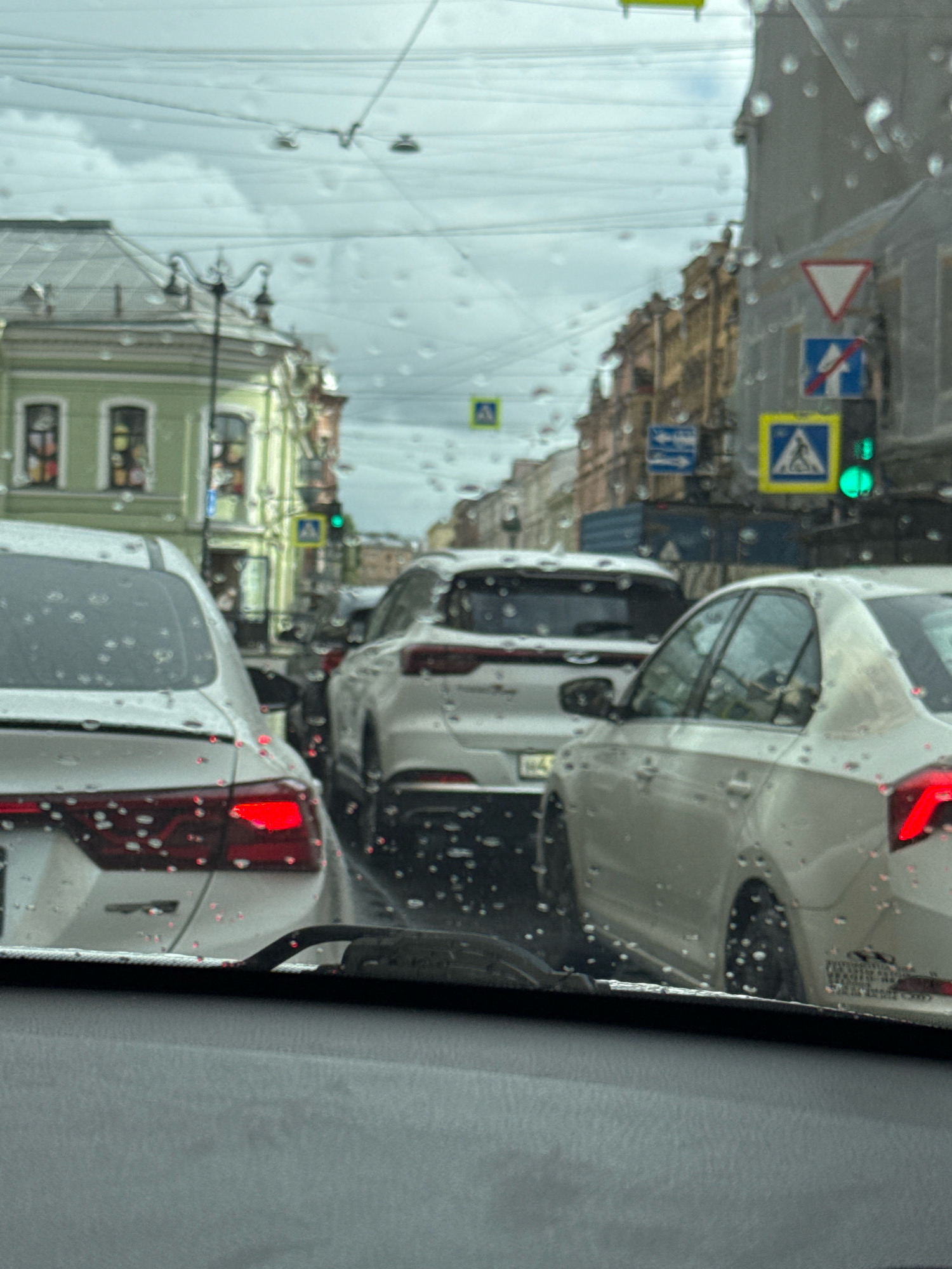 The image shows a view from inside a vehicle looking through a rain-streaked windshield at a street scene in Saint Petersburg, Russia. The street is lined with multi-story buildings, some with green facades and ornate details. Traffic is backed up, with several cars visible in the foreground and middle ground, including a white sedan with its brake lights illuminated and a white SUV. Traffic lights and road signs, including a pedestrian crossing sign and a yield sign, are visible. The sky is overcast and the weather appears to be rainy.