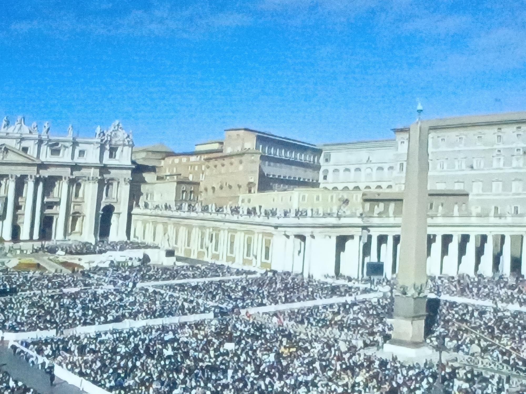 The image shows a large crowd of people gathered in St. Peter's Square in Vatican City. In the foreground, the crowd fills most of the square, with some people holding up signs or banners. In the middle ground, there is the Vatican Obelisk, surrounded by rows of columns that form the colonnade of the square. In the background, the facade of St. Peter's Basilica and other buildings within Vatican City can be seen under a clear blue sky.
The location in the city of Juan Griego, Venezuela is not visible in the image.