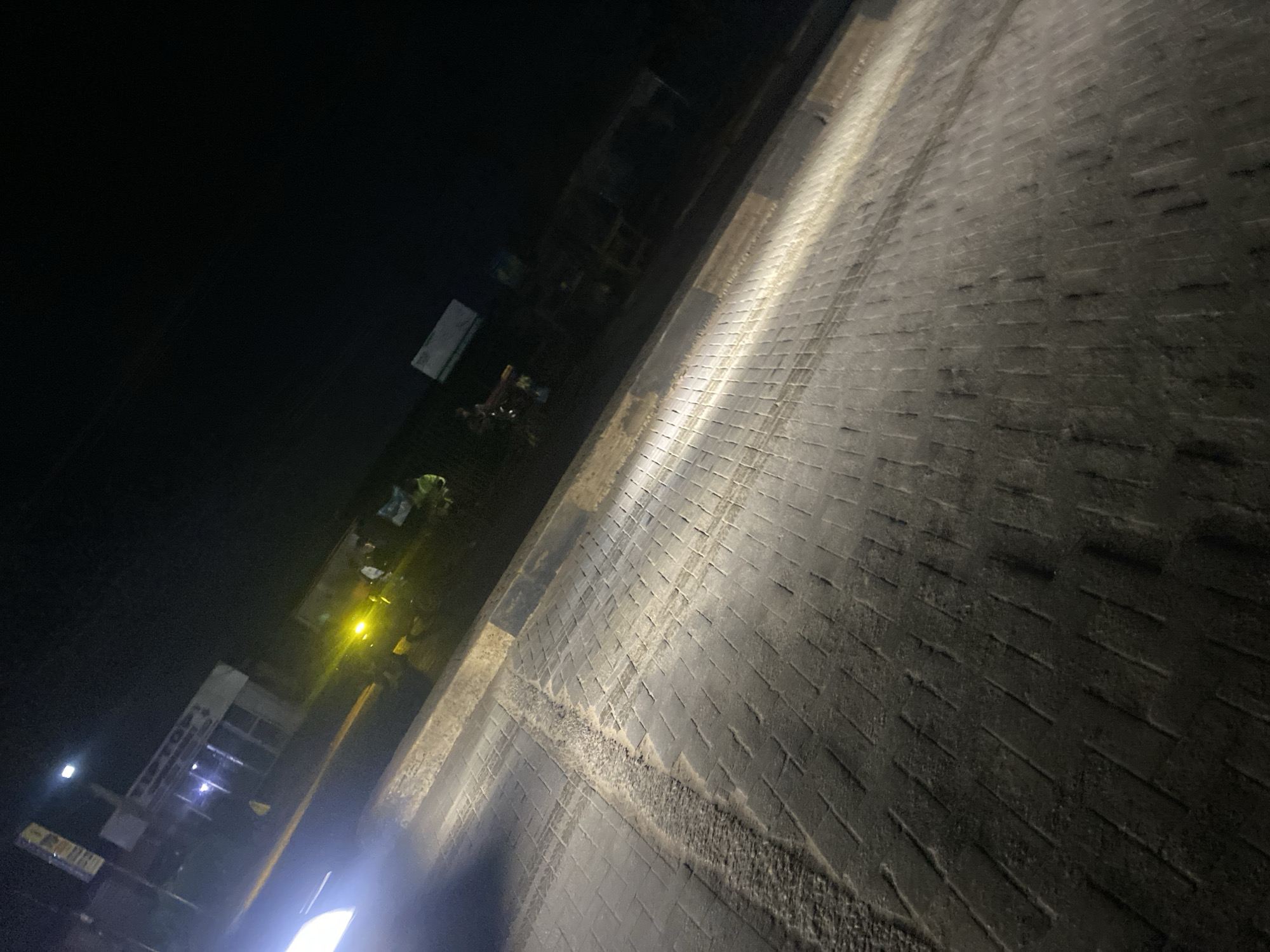 The image depicts a nighttime street scene in Ikorodu, Nigeria. The foreground is dominated by a paved road made of interlocking rectangular bricks, illuminated by a bright, diffuse white light originating from the lower-left, consistent with vehicle headlights. Tire marks are evident on the brick surface. A raised curb, marked with alternating black and yellow sections, runs along the right edge of the illuminated road, separating it from an unpaved, darker area.
In the midground, several indistinct human figures are visible on the right side of the road, gathered near multiple yellow light sources, which may belong to other vehicles or commercial establishments. On the left side of the road, a building with illuminated windows is present, featuring a sign with partially legible text that appears to include "SOFT." The background is dark, indicating a lack of ambient light from the sky. No clear actions or interactions among the individuals are discernible due to the low light conditions and distance.